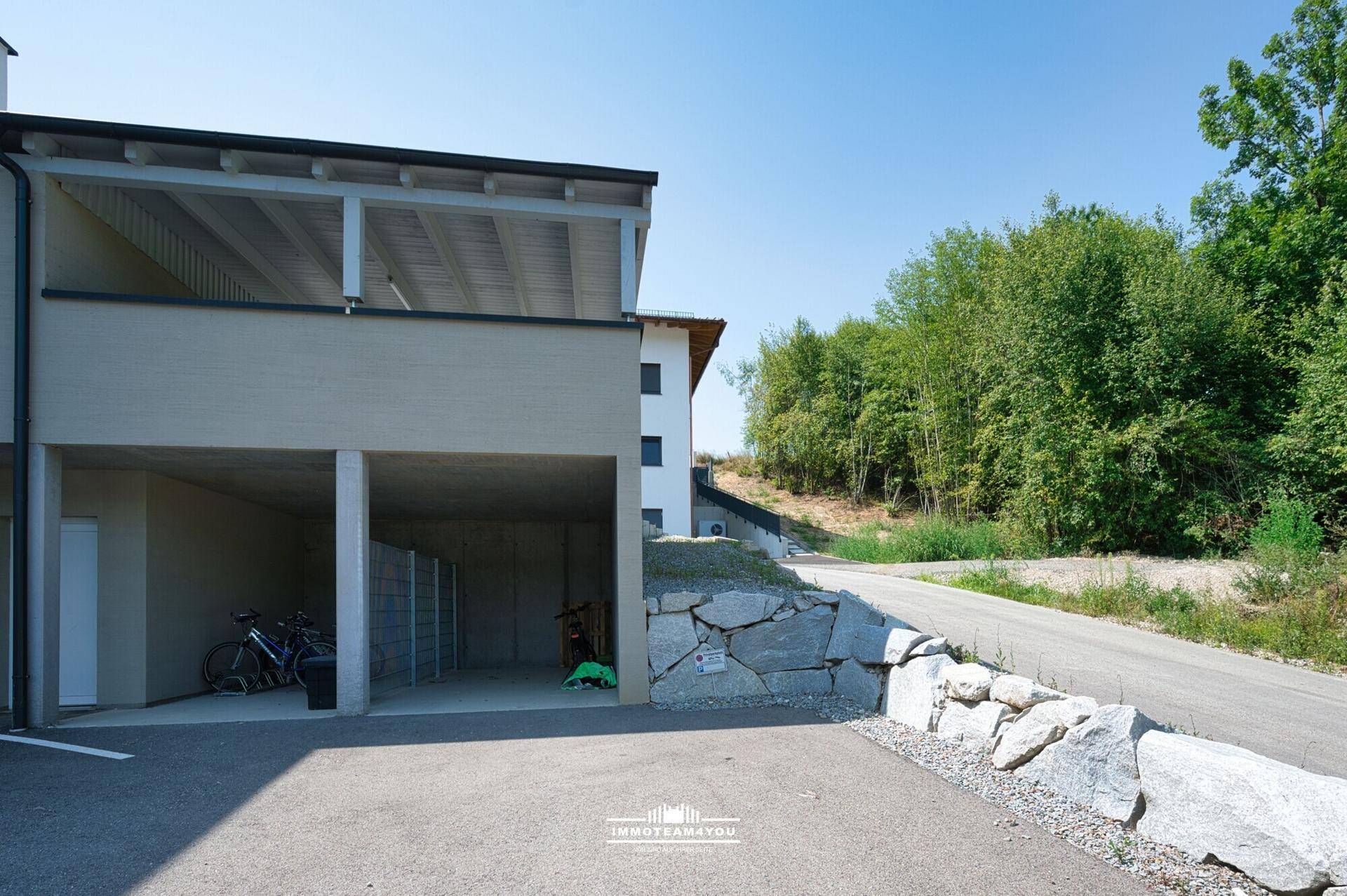 Überdachter Parkbereich neben dem Gebäude mit Zufahrt und Natursteinmauer.