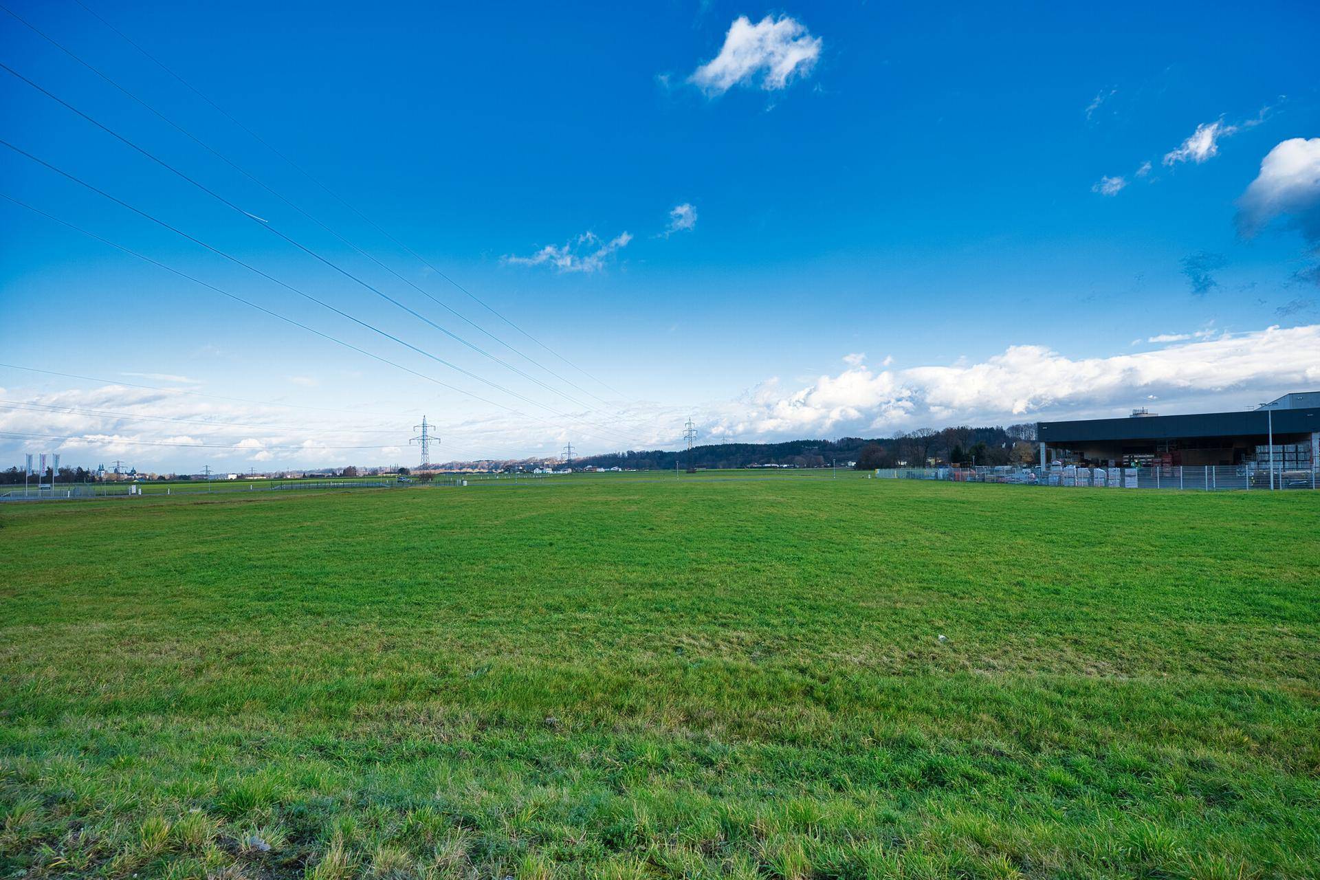 Weitläufige grüne Wiese unter blauem Himmel mit Stromleitungen und Gebäuden im Hintergrund.