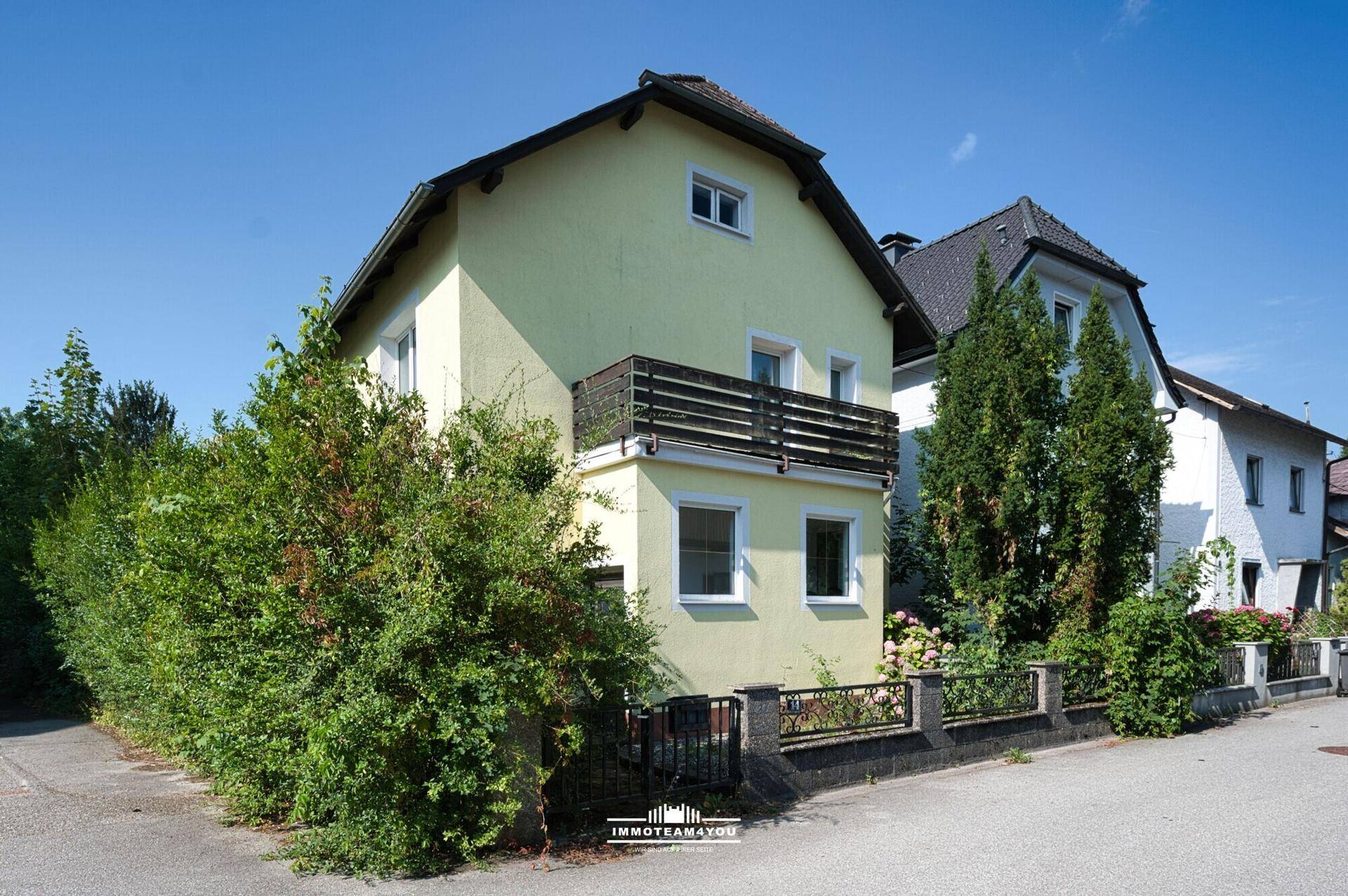 Seitenansicht des gelben Einfamilienhauses mit Balkon und umgebender grüner Hecke.
