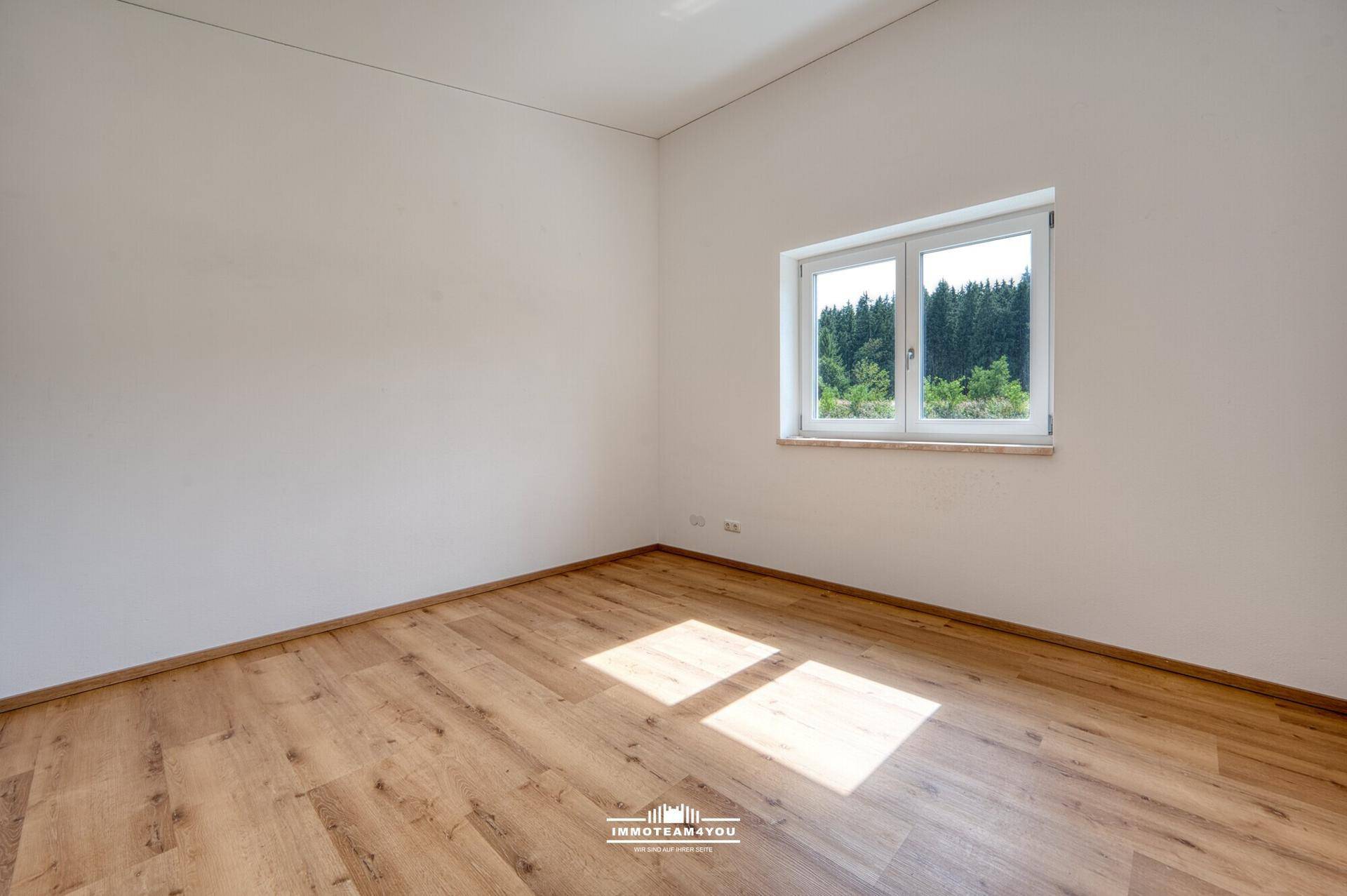 Leeres Schlafzimmer mit Holzboden, weißen Wänden und Fenster mit Blick ins Grüne.