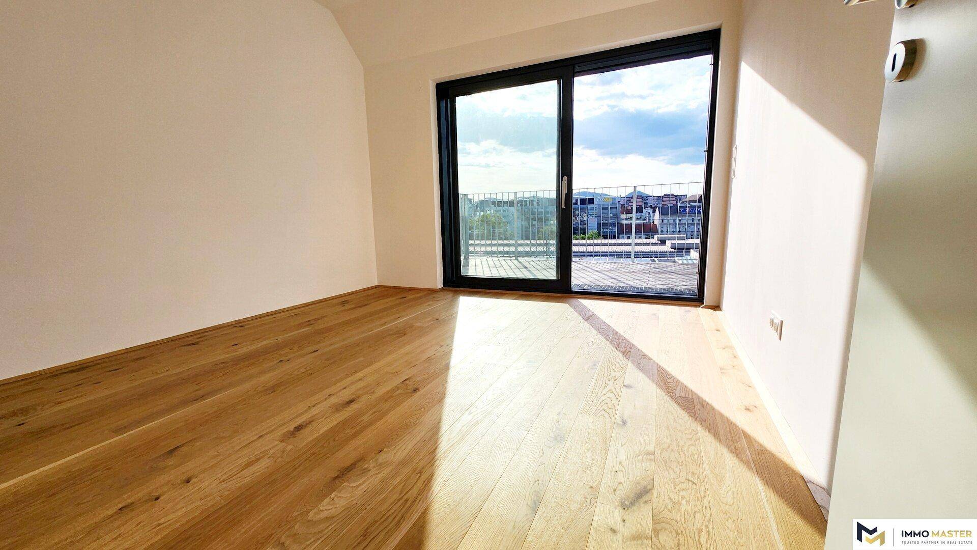 Helles Zimmer mit Holzboden und großer Glasschiebetür, die auf einen Balkon oder eine Terrasse führt.