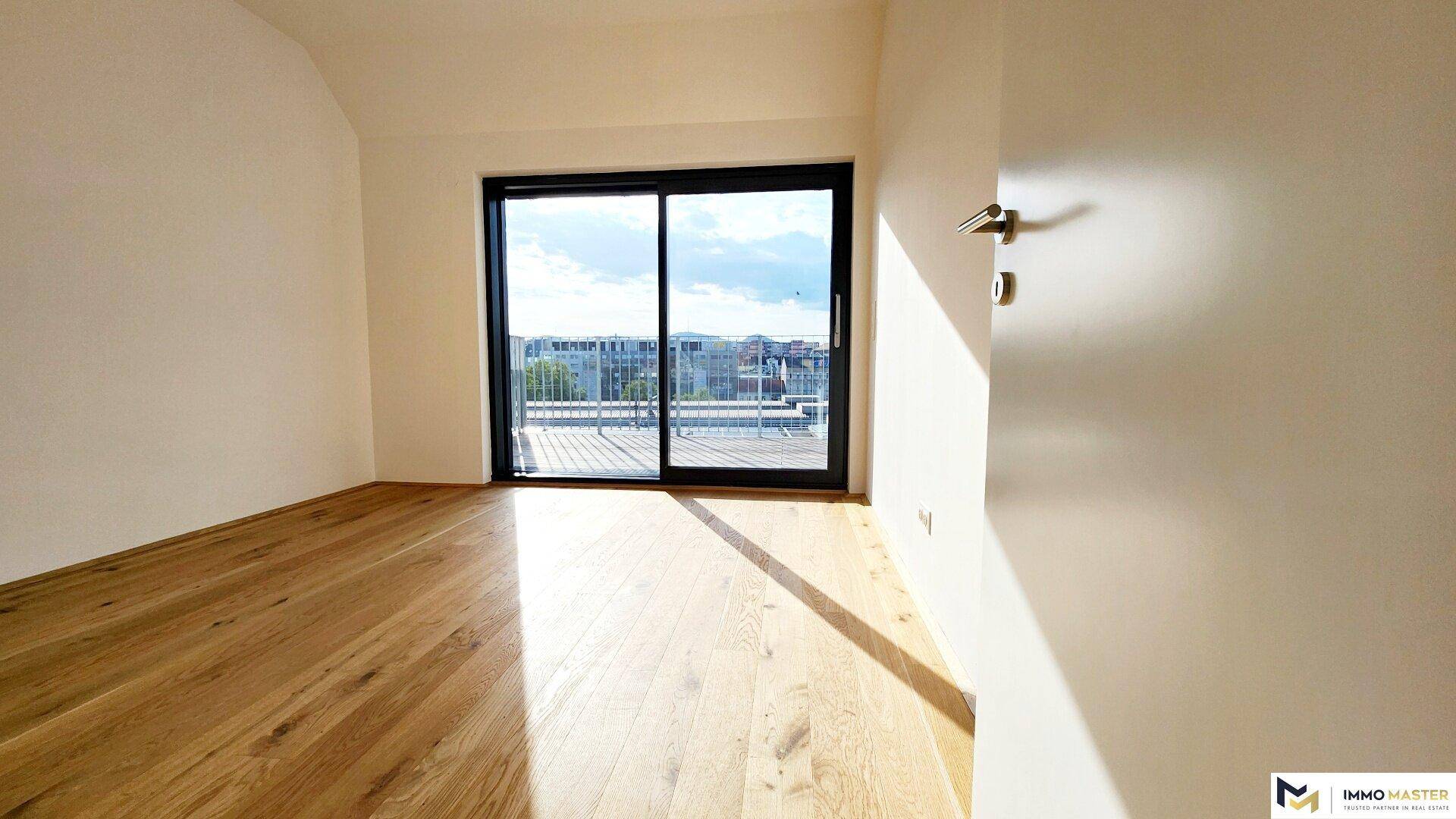 Helles Zimmer mit Holzboden und großer Glasschiebetür, die auf einen Balkon oder eine Terrasse führt.