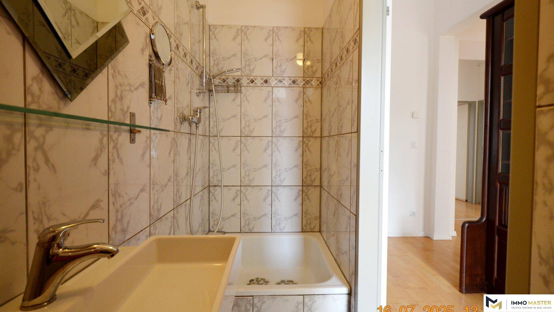 Badezimmer mit Duschwanne, Waschbecken und Spiegel, umgeben von hellen Marmorfliesen.