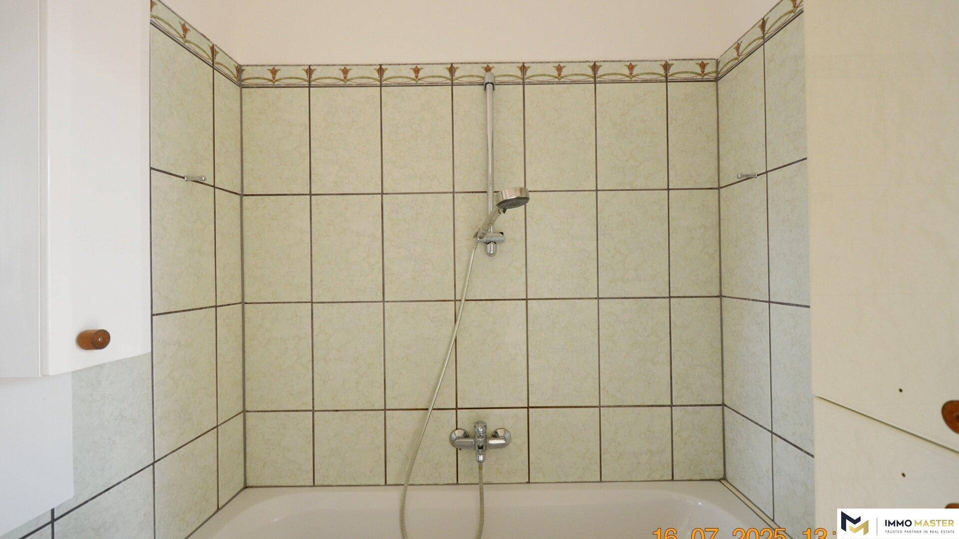 Badezimmer mit Badewanne und Duscharmatur, umgeben von hellen Fliesen mit Bordüre.