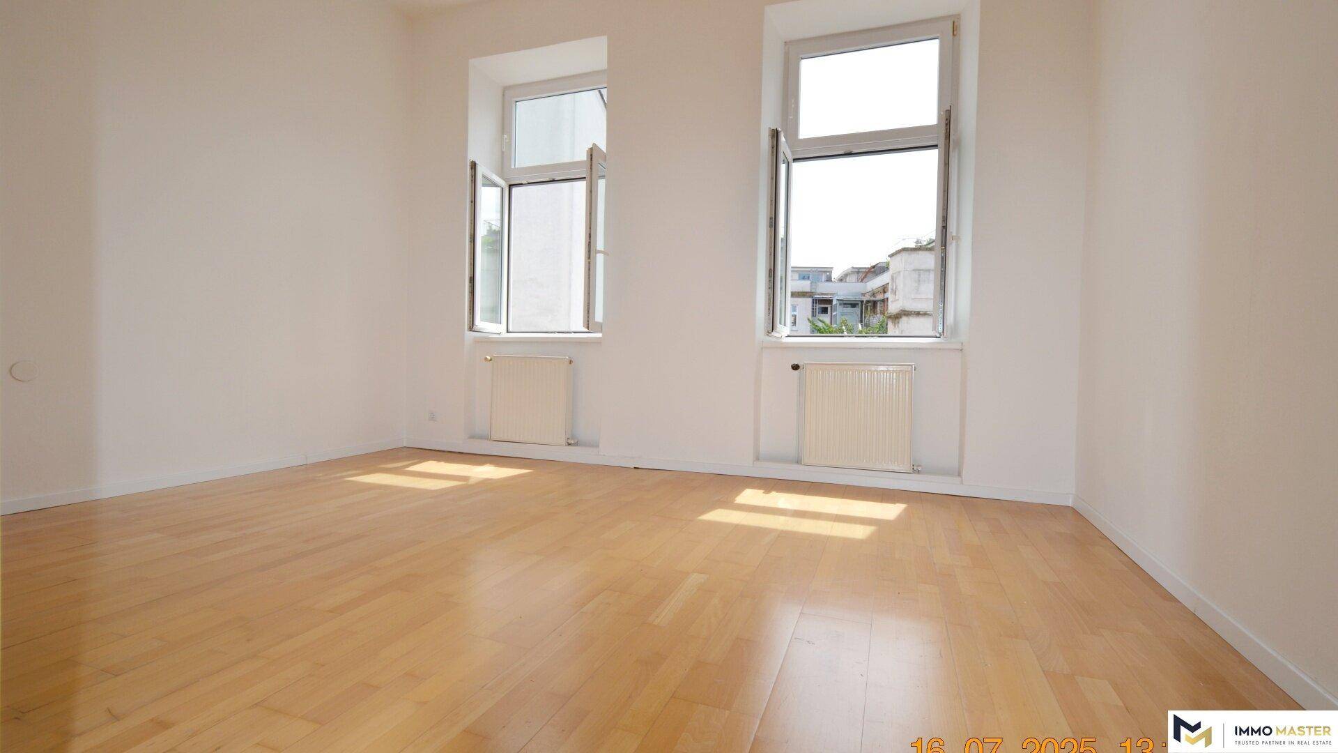 Heller Wohnraum mit Holzboden und zwei großen Fenstern, die viel Tageslicht hereinlassen.