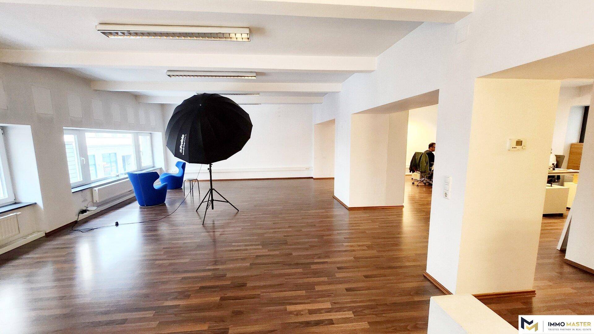 Offener Bürobereich mit Holzboden, großen Fenstern und einem Blick in angrenzende Arbeitsbereiche.