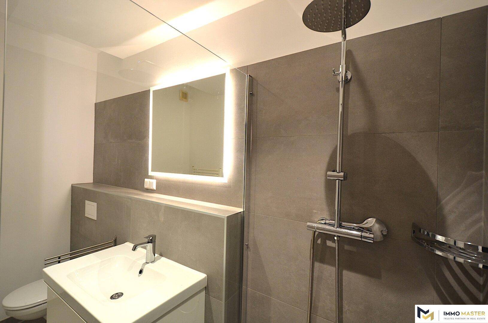 Modernes Badezimmer mit grauen Fliesen, begehbarer Dusche und beleuchtetem Spiegel.