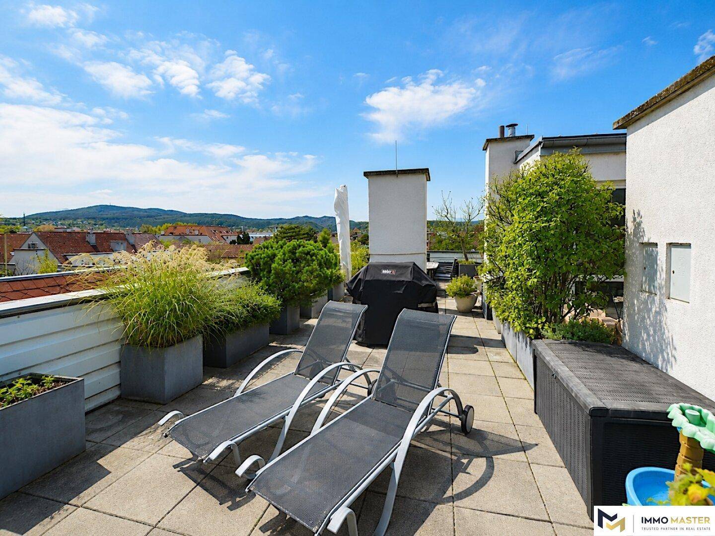 Sonniger Dachgarten mit Liegestühlen, Grillbereich und weitem Blick auf die umliegende Landschaft.