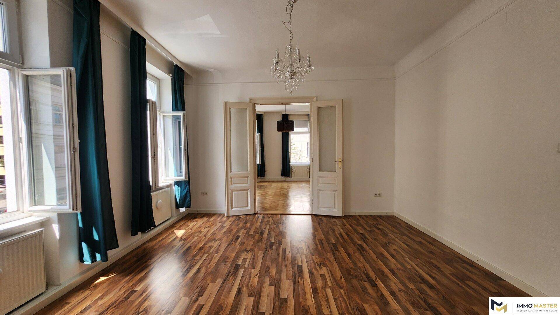 Leeres Wohnzimmer mit Holzboden, hohen Decken und Flügeltüren zum nächsten Raum.