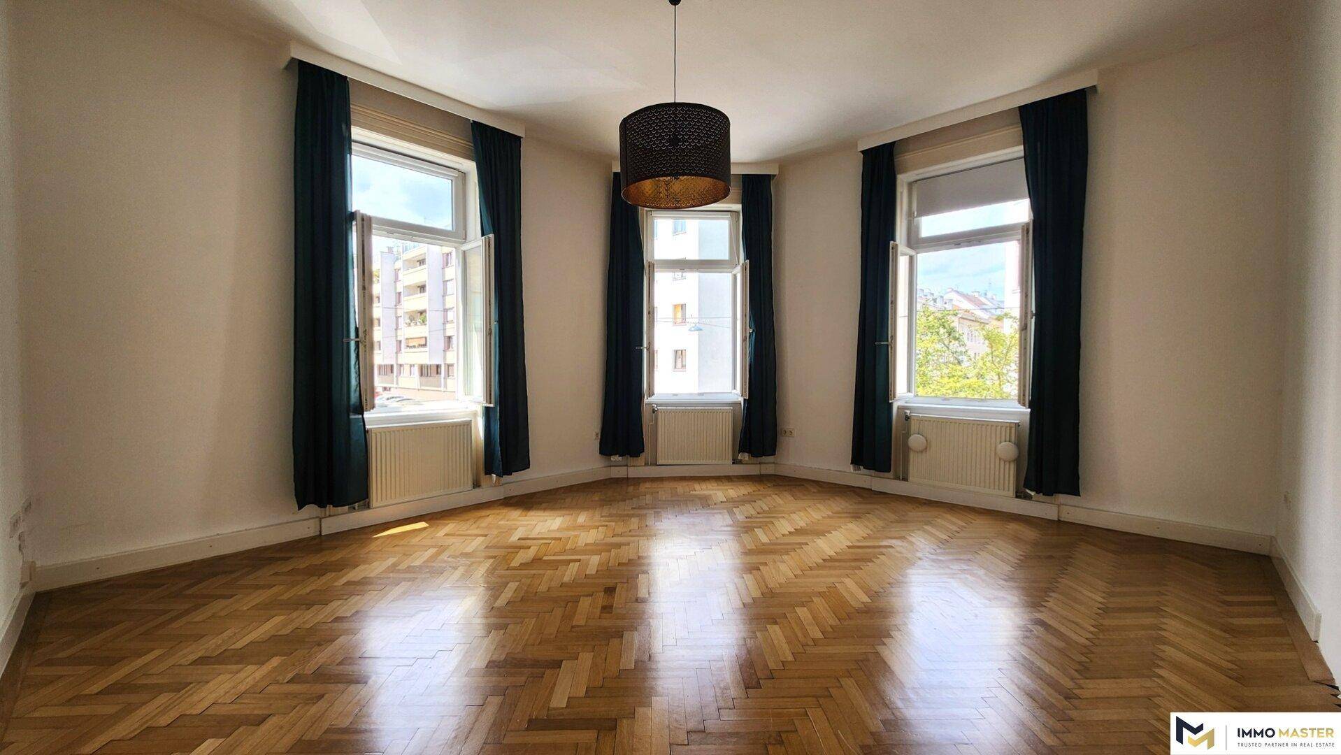 Geräumiges, leeres Wohnzimmer mit Fischgrätparkett und drei großen Fenstern.