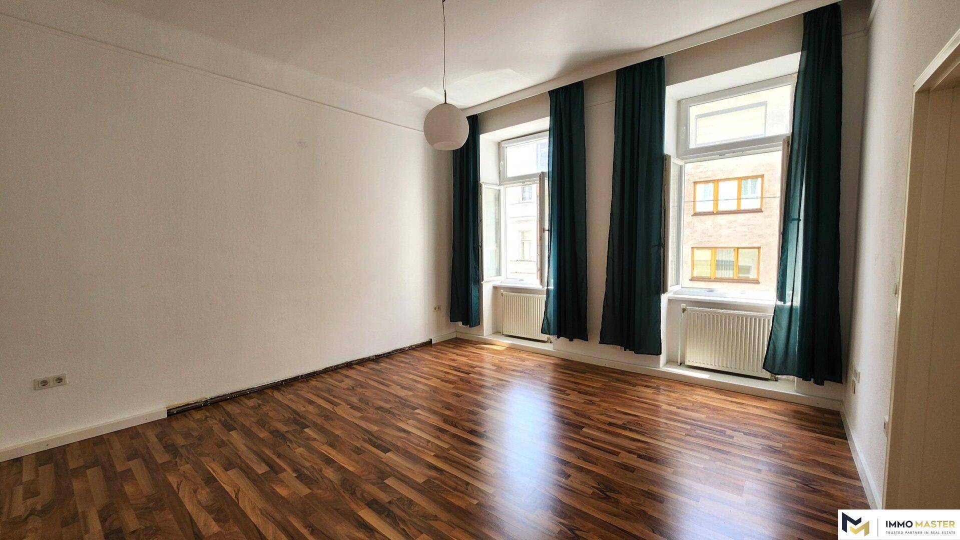 Leeres Schlafzimmer mit Holzboden, hohen Decken und zwei Fenstern mit dunklen Vorhängen.