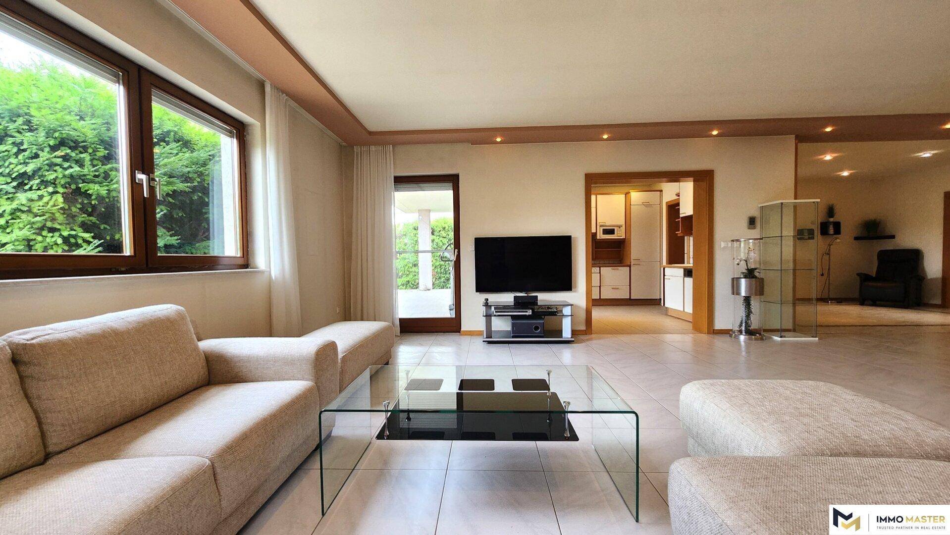 Geräumiges Wohnzimmer mit beigem Sofa, Glastisch und großen Fenstern mit Blick ins Grüne.