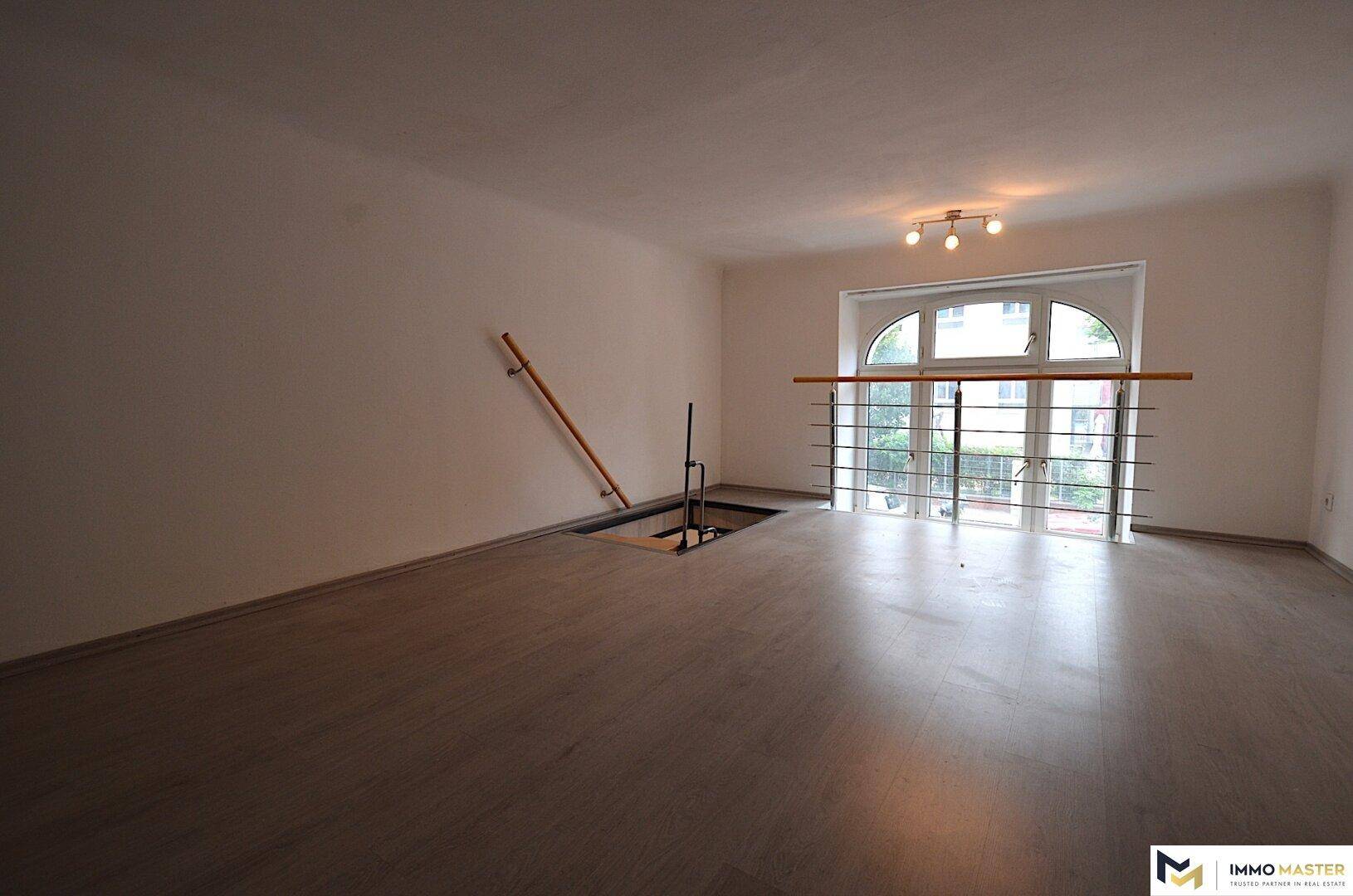 Geräumiger Wohnbereich mit dunklem Laminatboden, einem Geländer und einem großen Bogenfenster mit Balkonzugang.