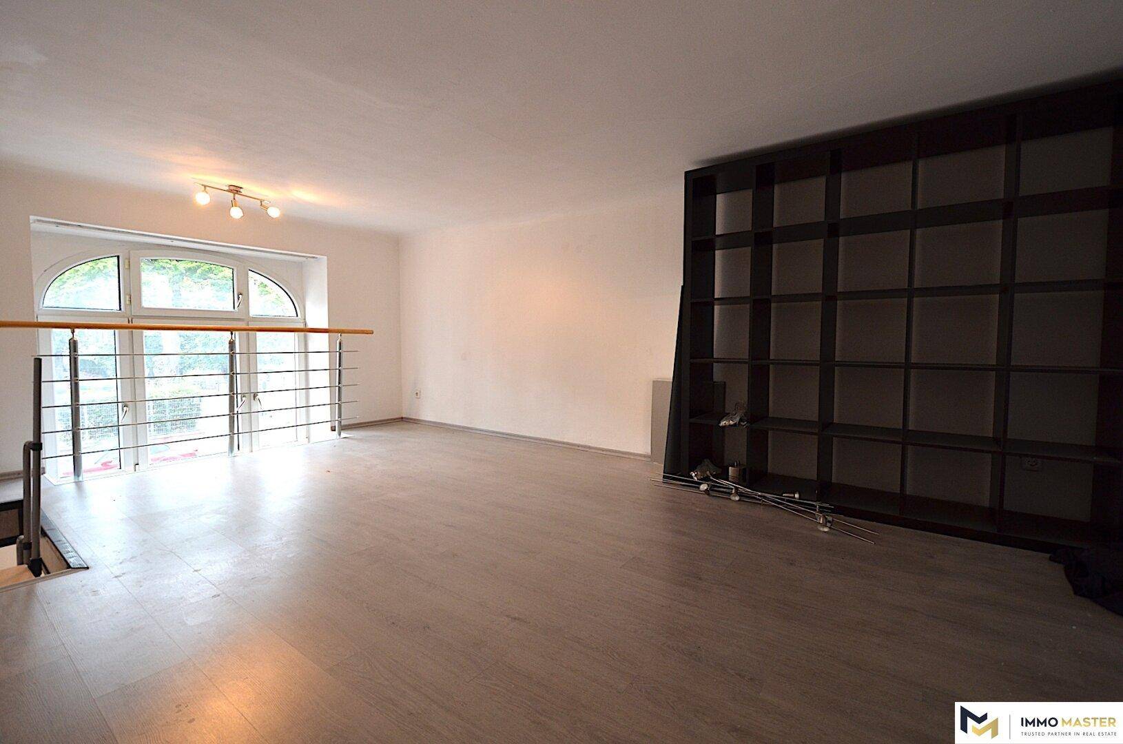 Heller Wohnraum mit dunklem Laminatboden, einem großen Bogenfenster und einem praktischen eingebauten Regal.