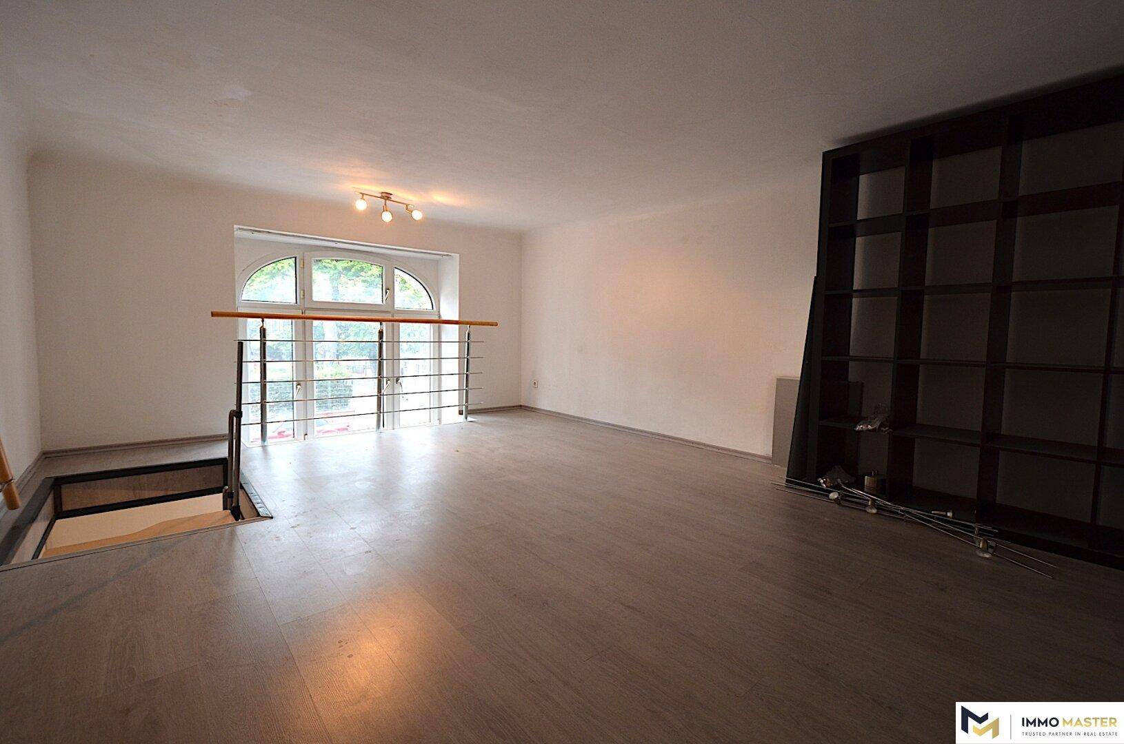 Geräumiger Wohnbereich mit Laminatboden, einem großen Bogenfenster und einer sichtbaren Treppenöffnung.
