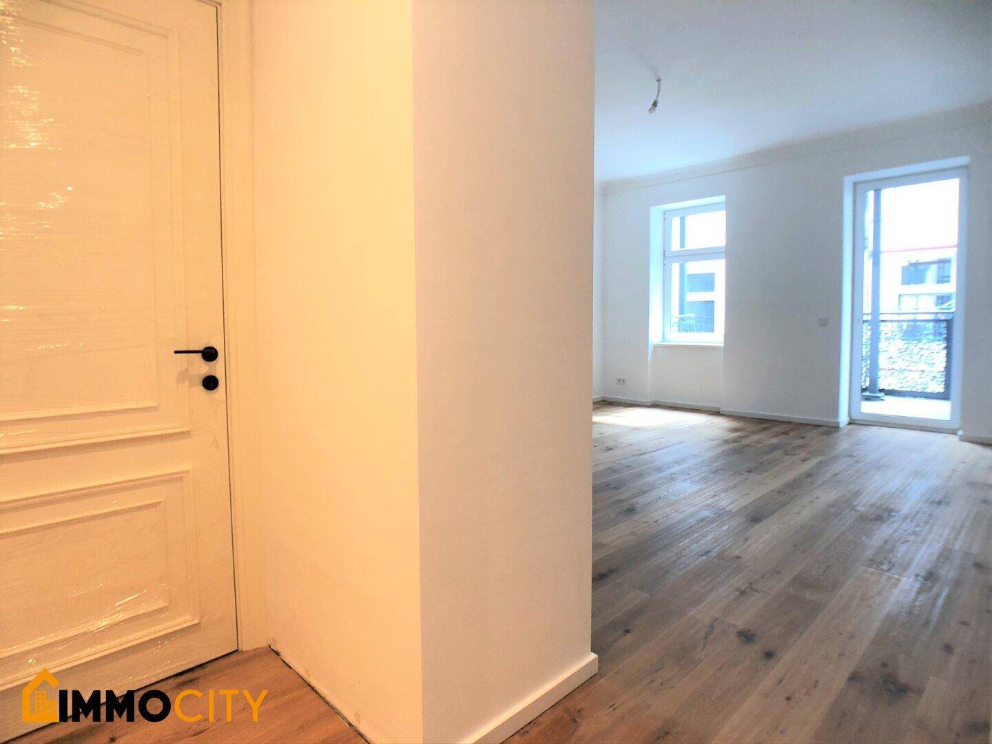 Blick vom Eingangsbereich in ein helles Zimmer mit Holzboden und Zugang zu einem Balkon.
