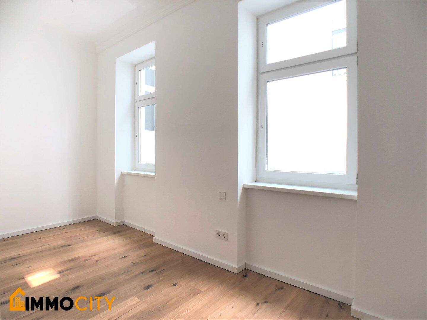 Helles Zimmer mit zwei großen Fenstern und Holzboden, ideal als Schlaf- oder Arbeitszimmer.