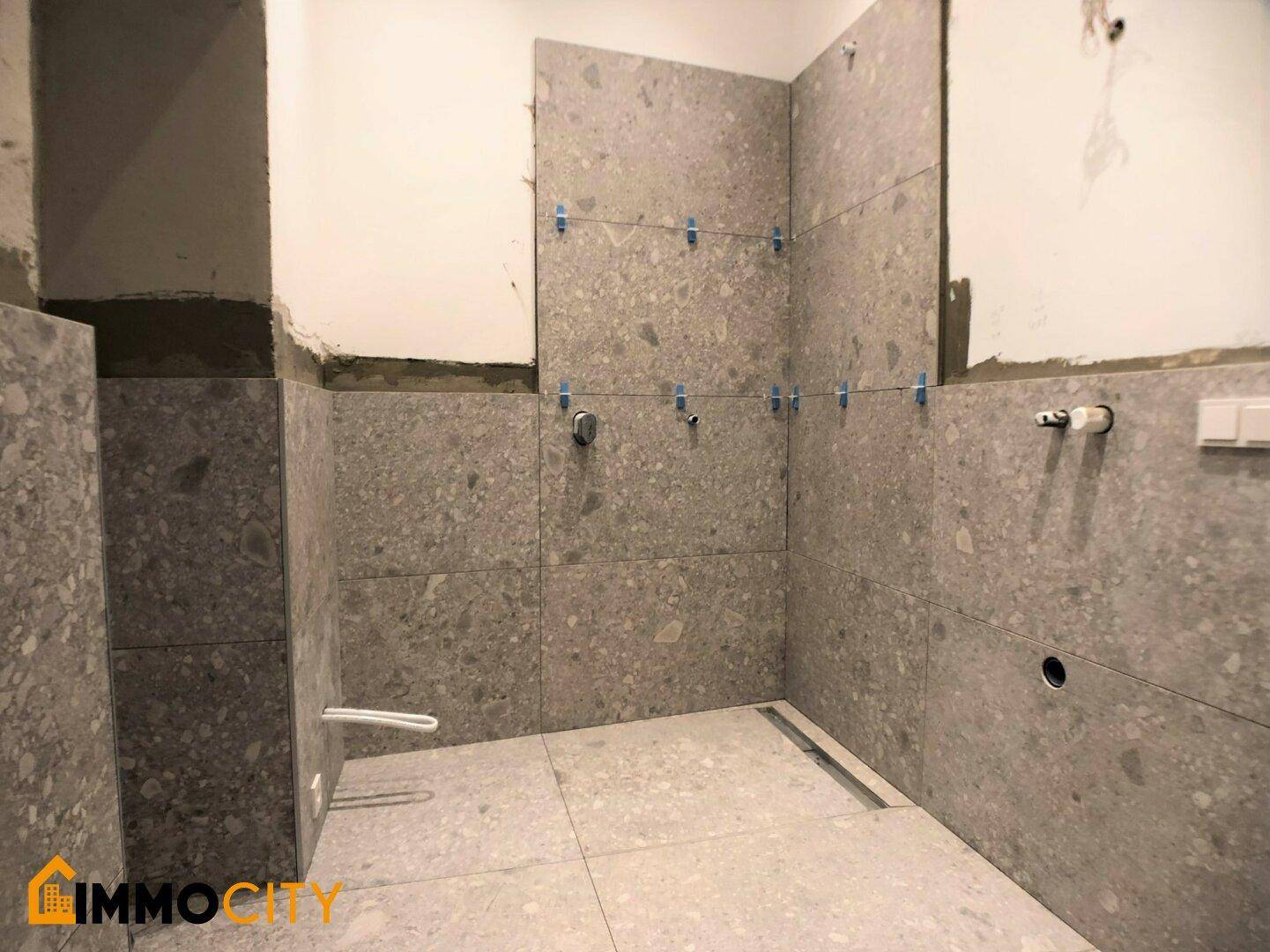 Unfertiges Badezimmer mit grauen Fliesen und vorbereiteten Installationen für Armaturen.