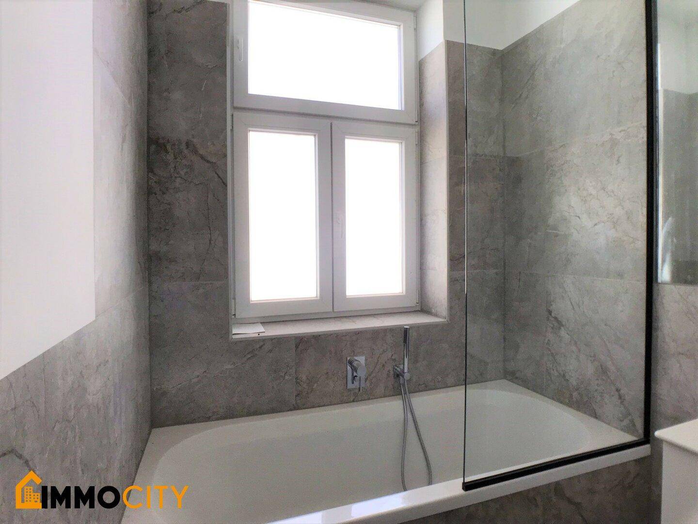 Modernes Badezimmer mit Badewanne und Fenster, umgeben von grauen Marmorfliesen an den Wänden.