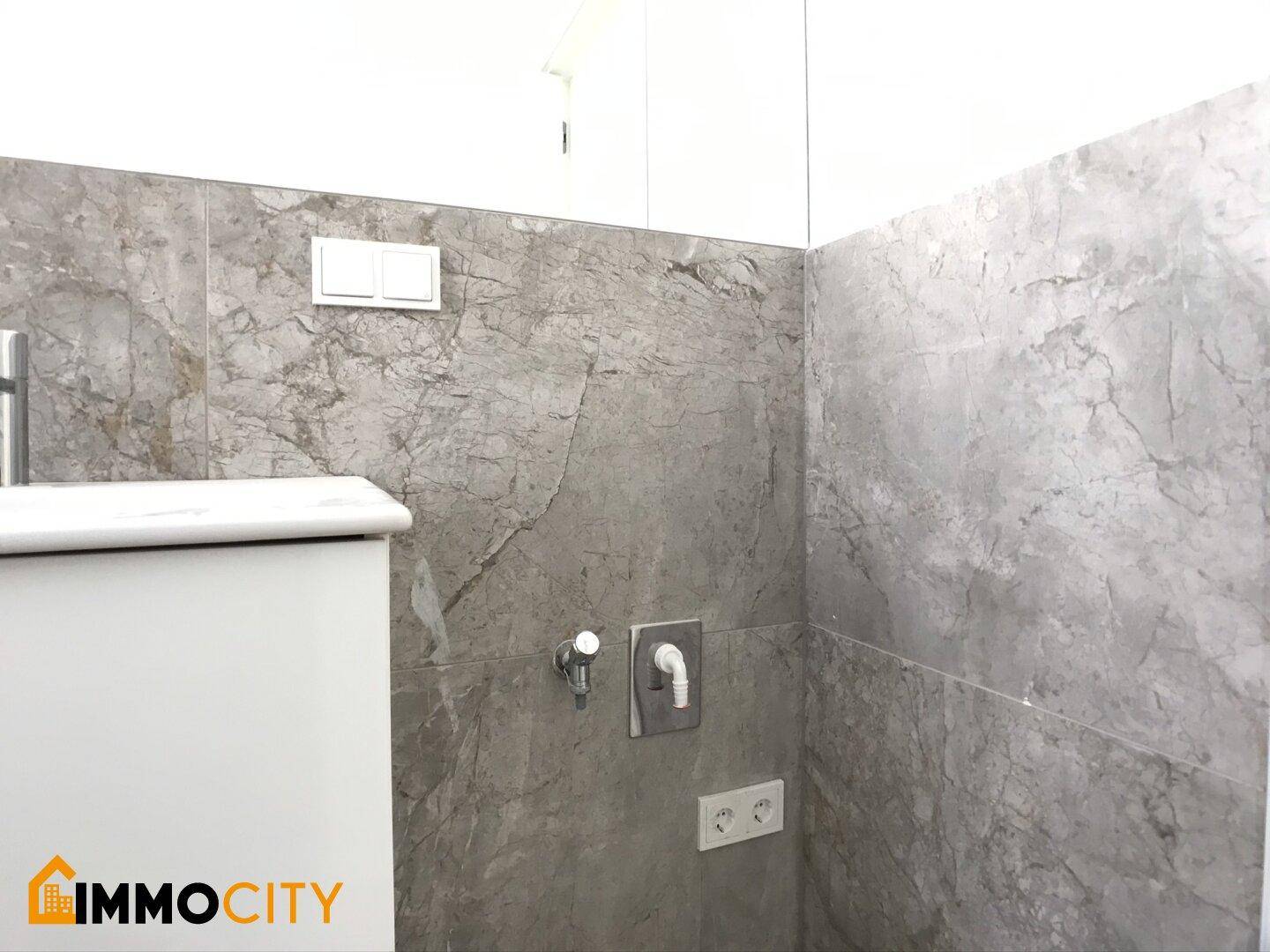 Detailansicht der grauen Fliesenwand im Badezimmer mit Armatur und Steckdose.