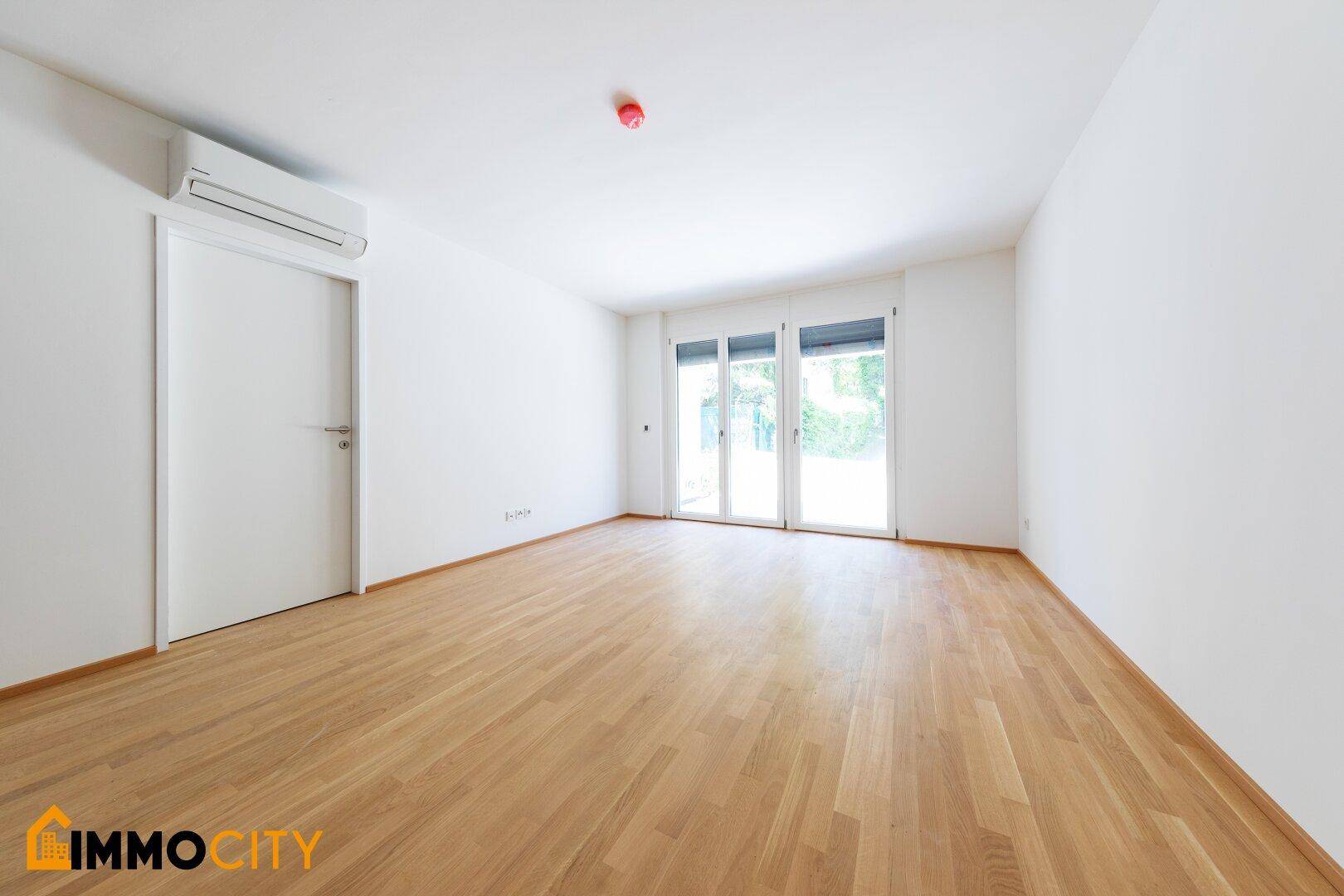 Lichtdurchfluteter Raum mit Parkettboden, Klimaanlage und Zugang zu einem Balkon oder Terrasse.