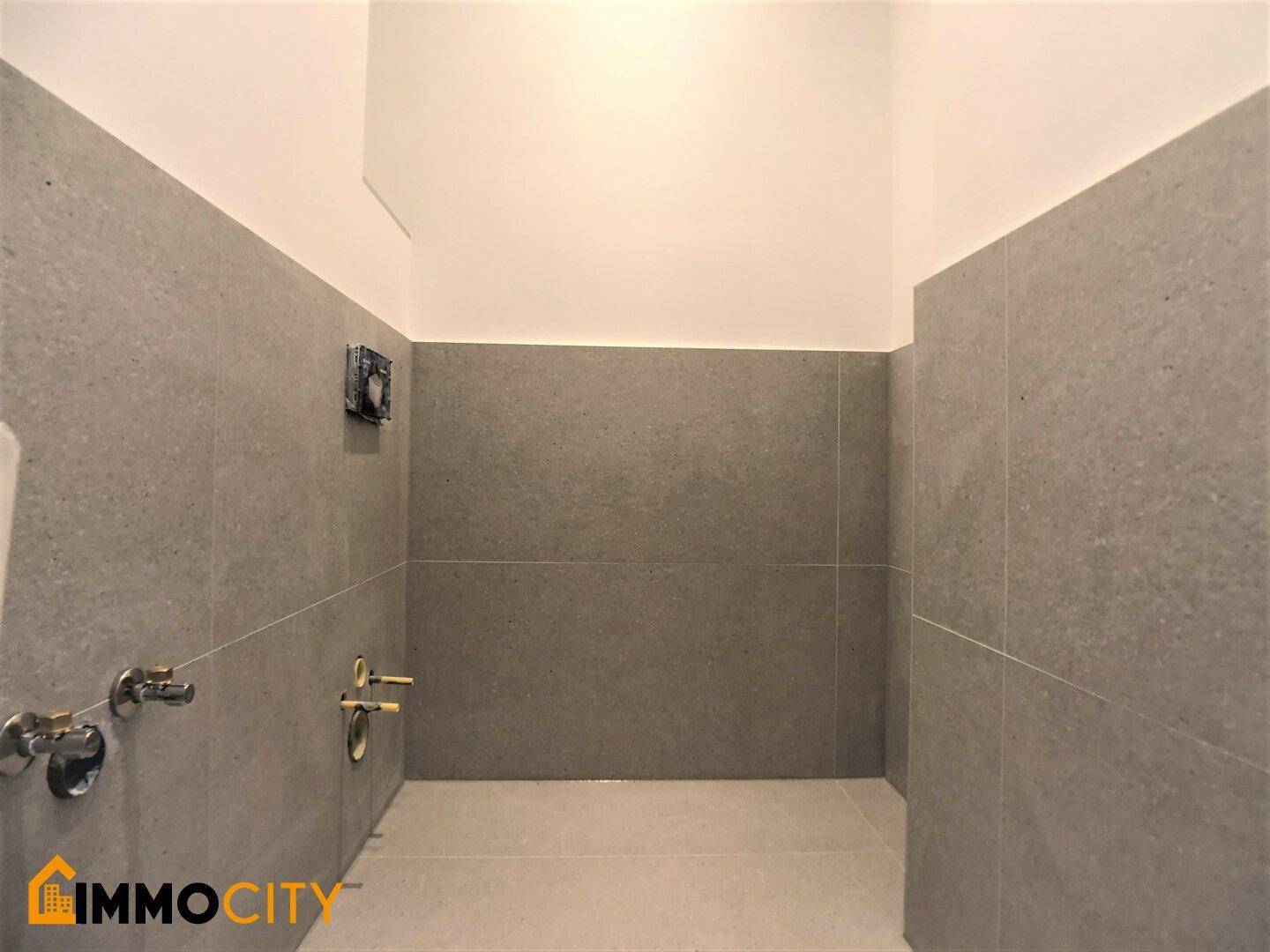Unfertiges Badezimmer mit grauen Wand- und Bodenfliesen sowie vorbereiteten Anschlüssen für Sanitäranlagen.