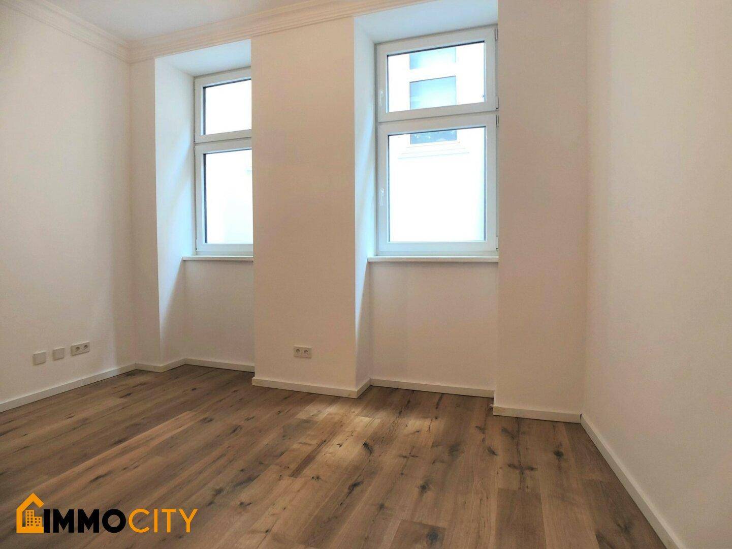 Geräumiges Zimmer mit neuem Holzboden und zwei großen Fenstern, bietet viel Tageslicht.