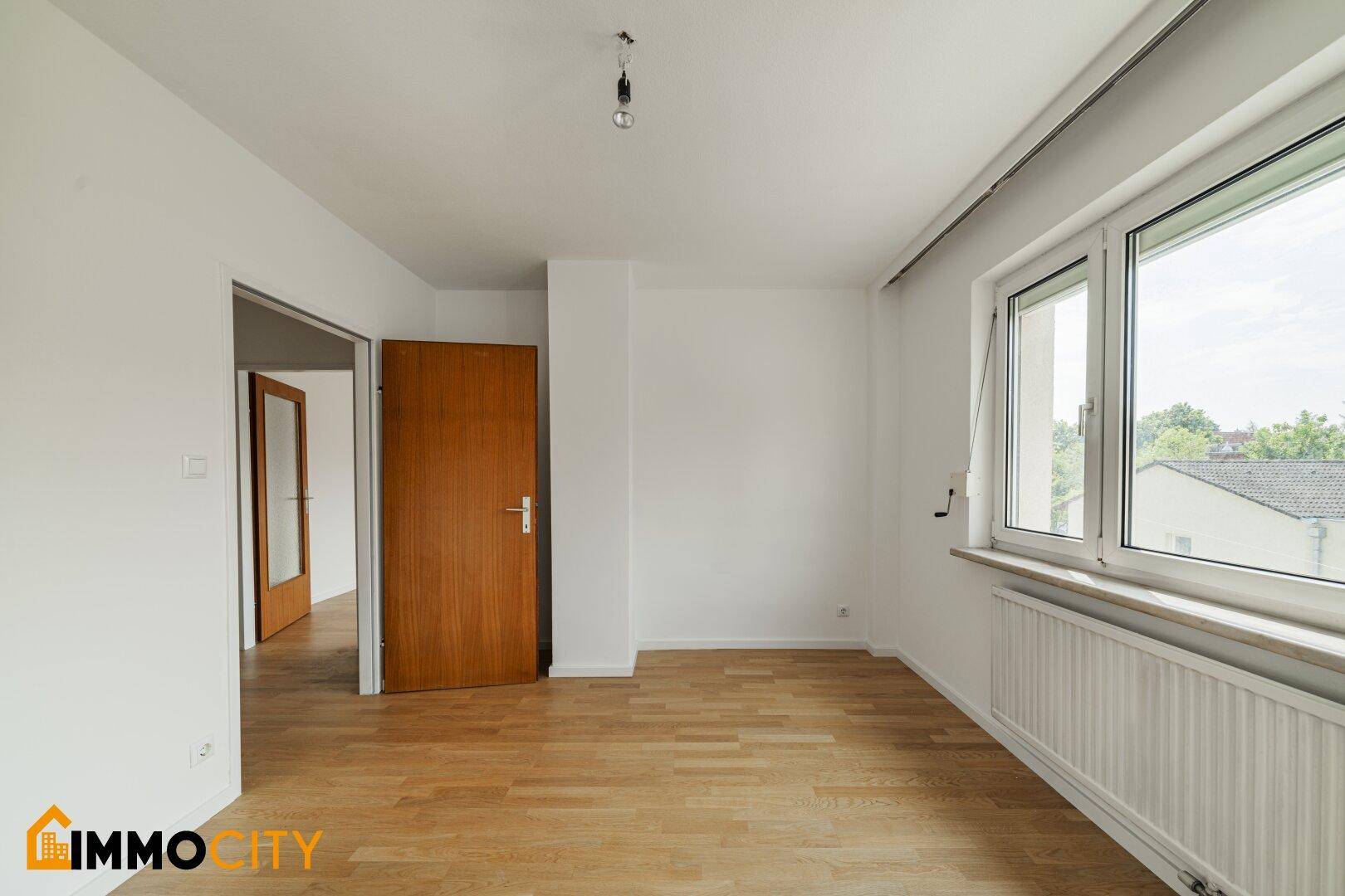 Geräumiges Zimmer mit Parkettboden und Fenster, das viel Tageslicht hereinlässt und einen Blick nach draußen bietet.