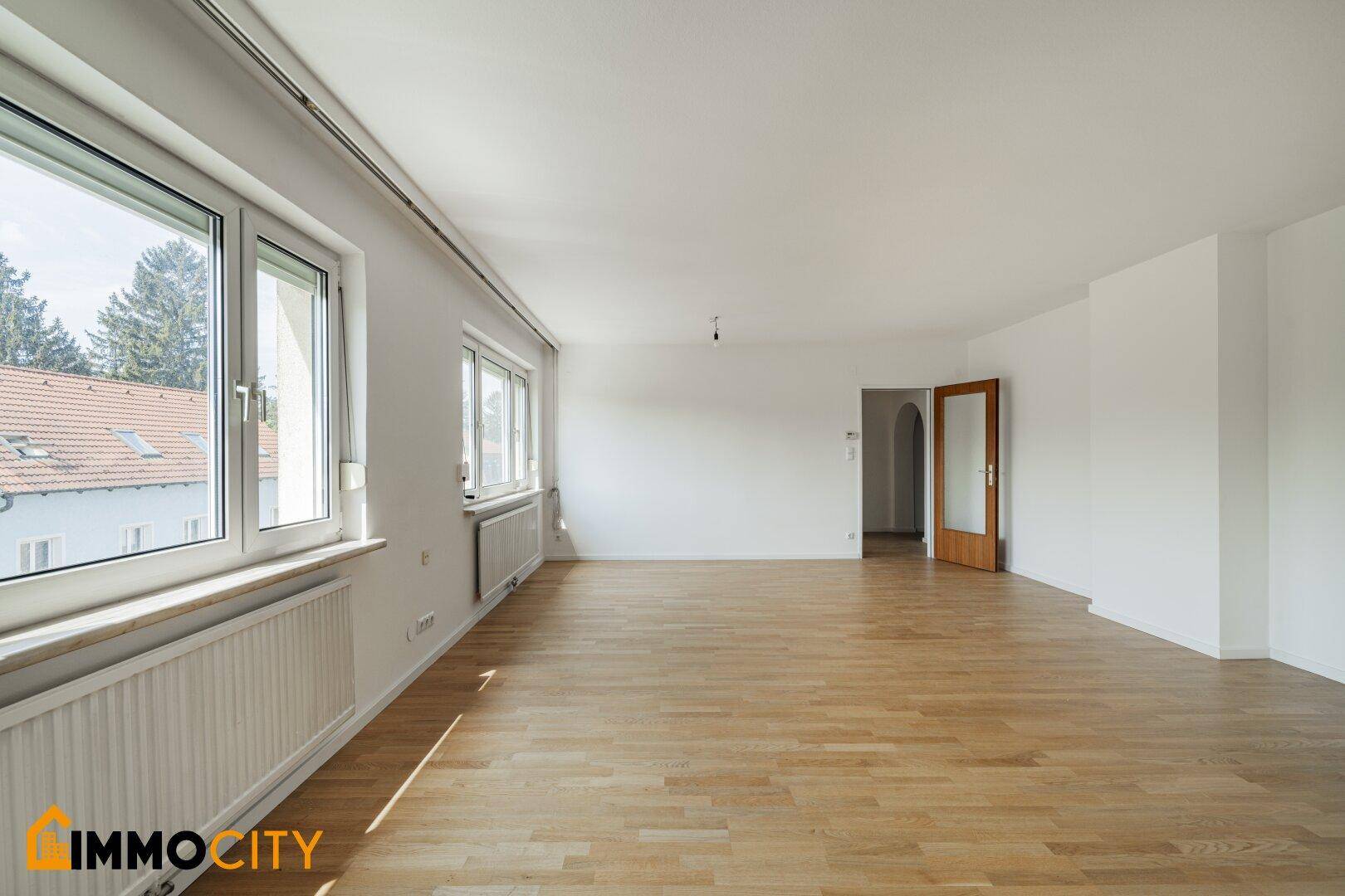 Langes, helles Wohnzimmer mit Parkettboden und einer Fensterfront, die viel Licht spendet.