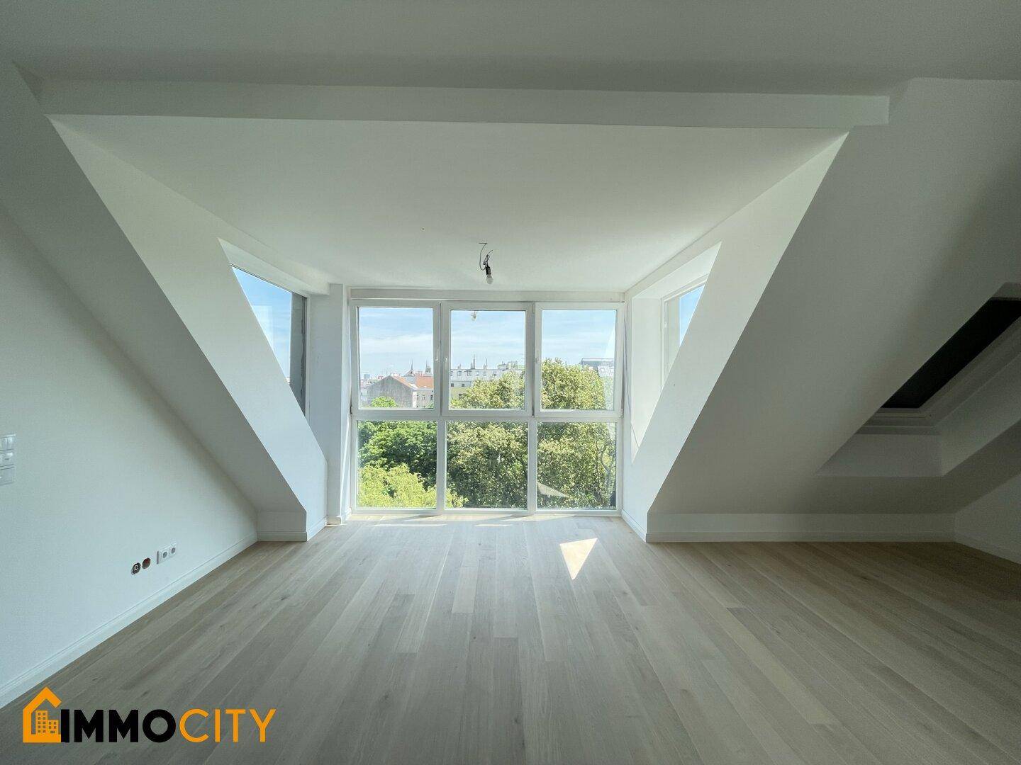 Helles Zimmer im Dachgeschoss mit großen Fenstern, Holzboden und schrägen Wänden.