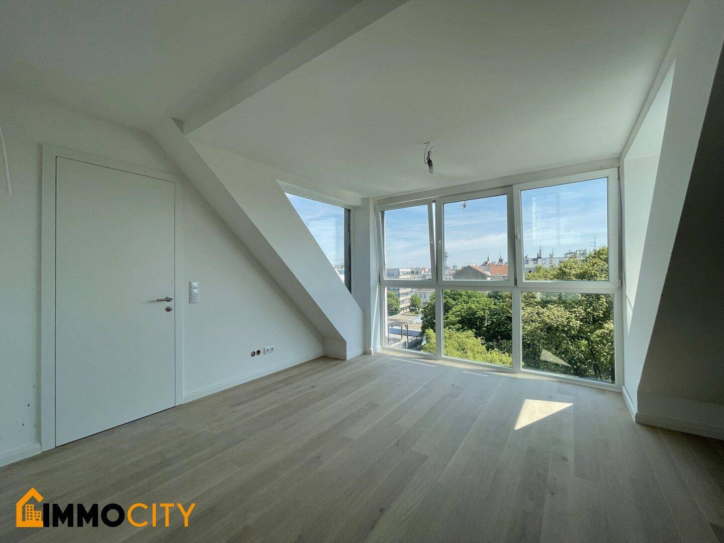 Helles Zimmer im Dachgeschoss mit großen Fenstern, Holzboden und schrägen Wänden.