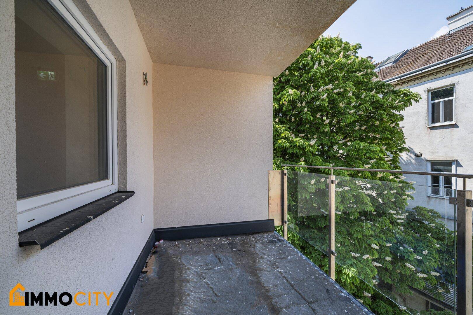 Kleiner Balkon mit Glasbrüstung und Blick auf grüne Bäume.