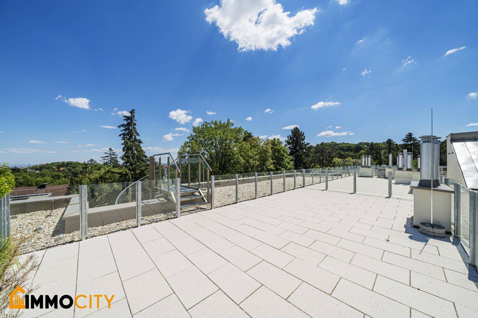 Weitläufige Dachterrasse mit hellem Fliesenboden und Glasgeländer, umgeben von viel Grün.