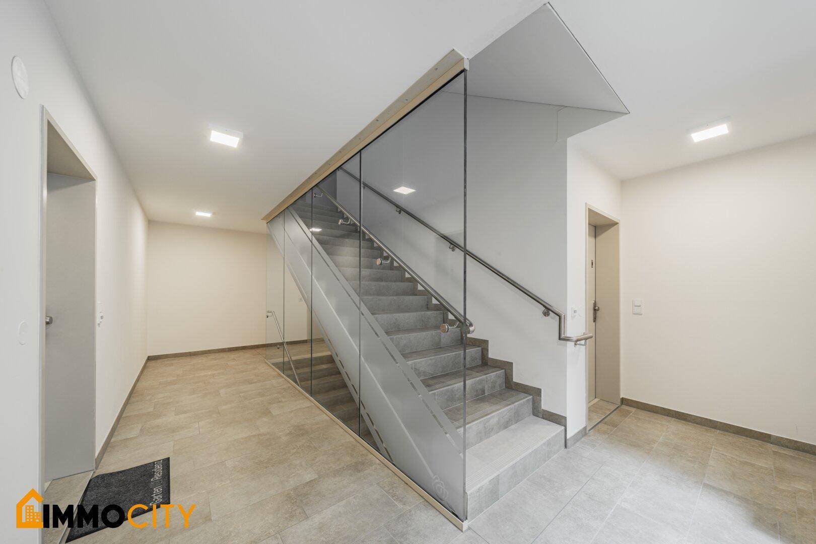 Heller Eingangsbereich mit moderner Treppe hinter Glas und Zugang zum Aufzug.