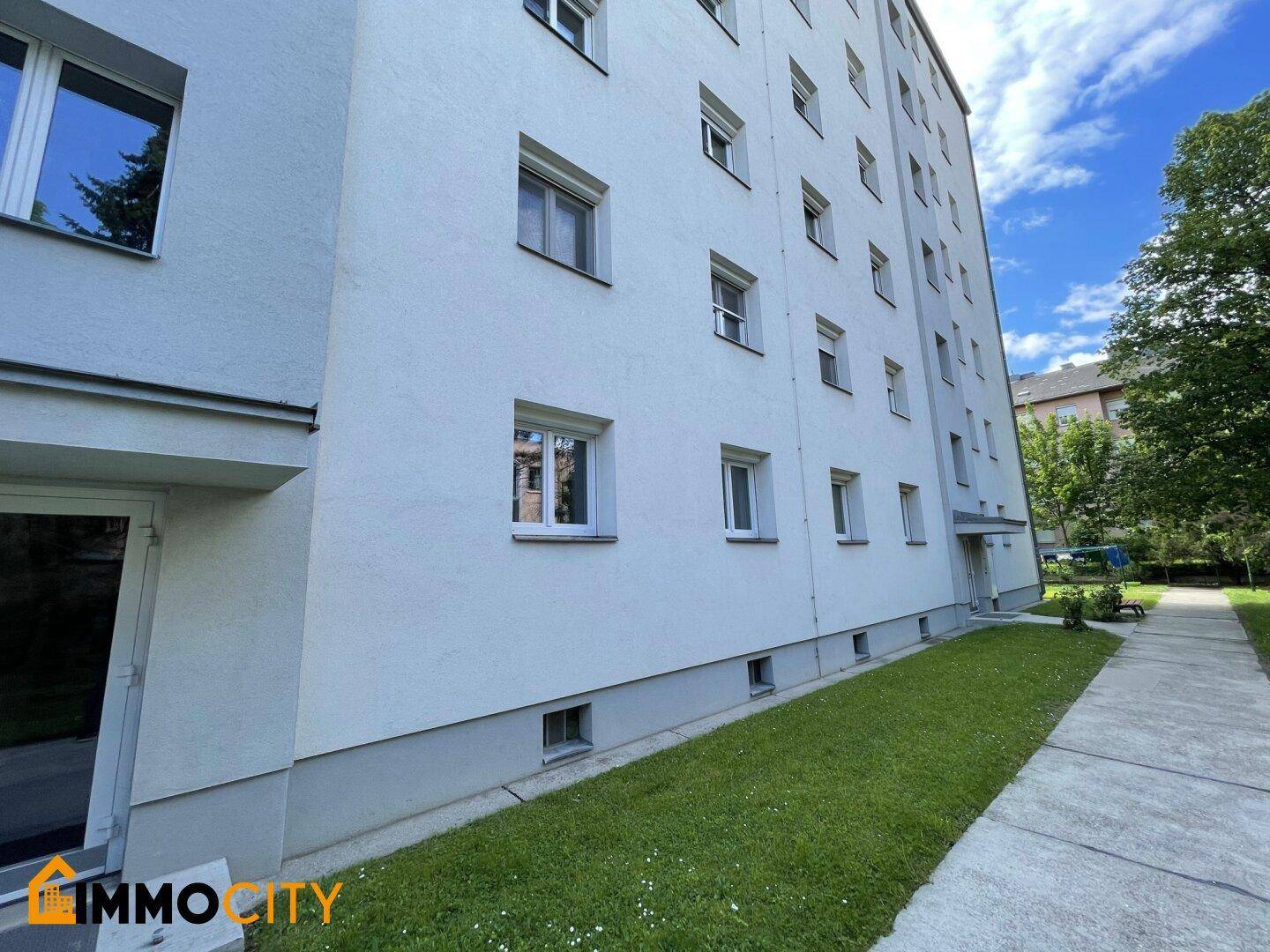 Seitenansicht eines Mehrfamilienhauses mit weißer Fassade und Zugang zum Gartenbereich.