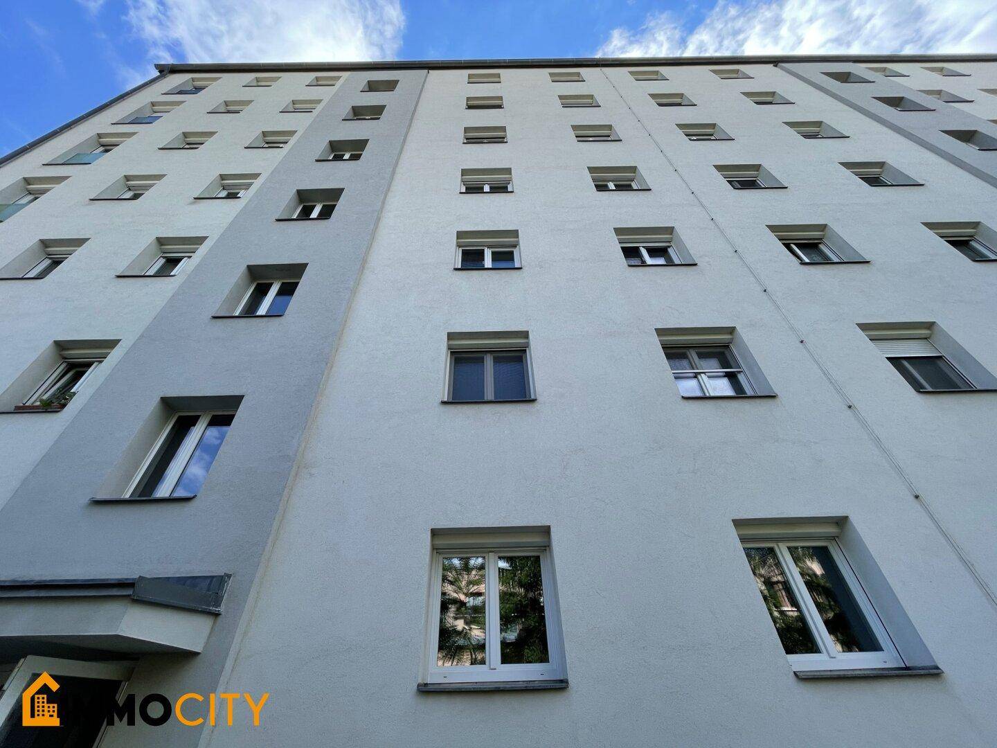 Hohe Fassade eines Mehrfamilienhauses mit vielen Fenstern unter blauem Himmel.