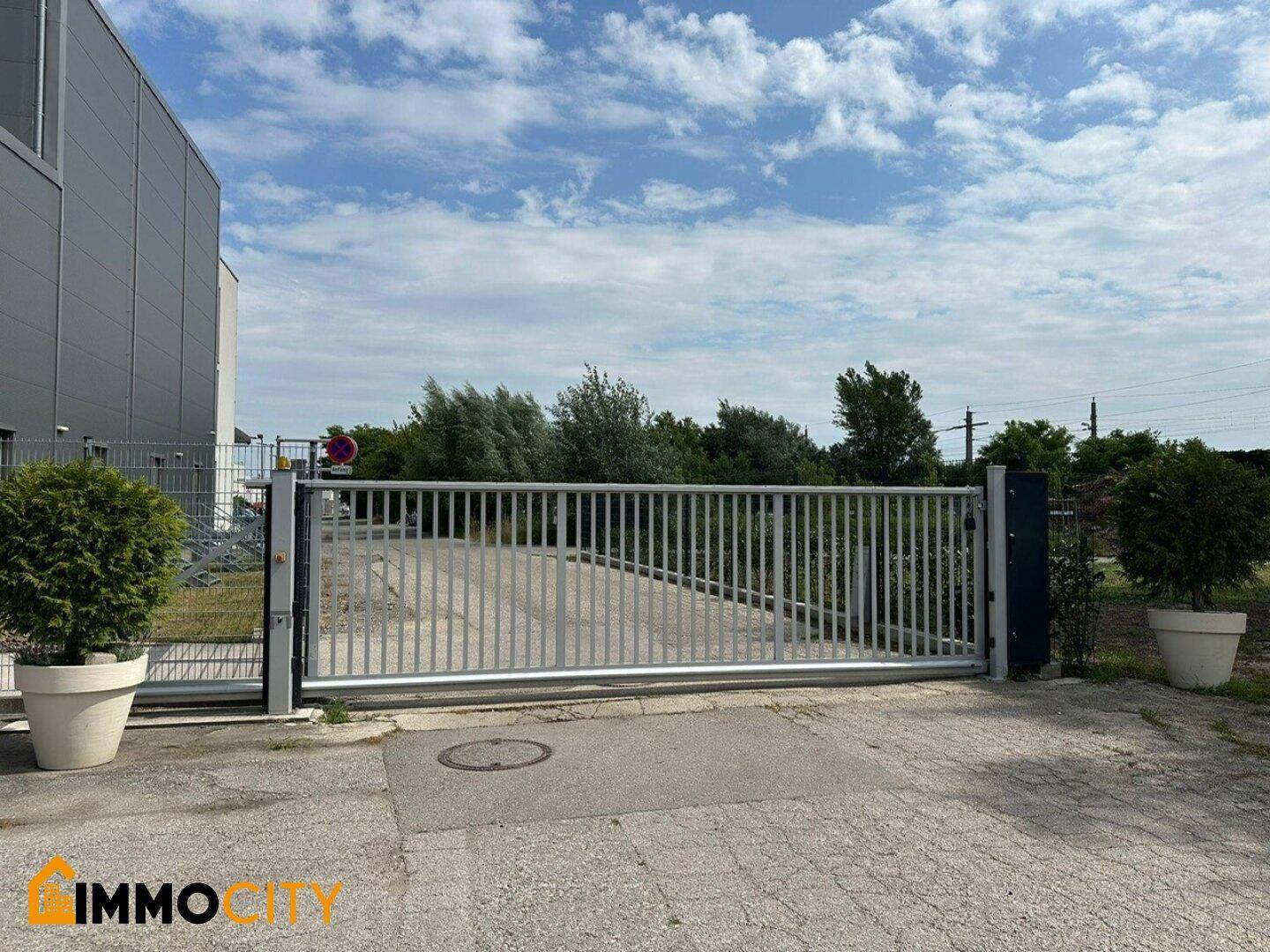 Großes Metalltor als Zufahrt zum Grundstück, flankiert von dekorativen Topfpflanzen unter bewölktem Himmel.