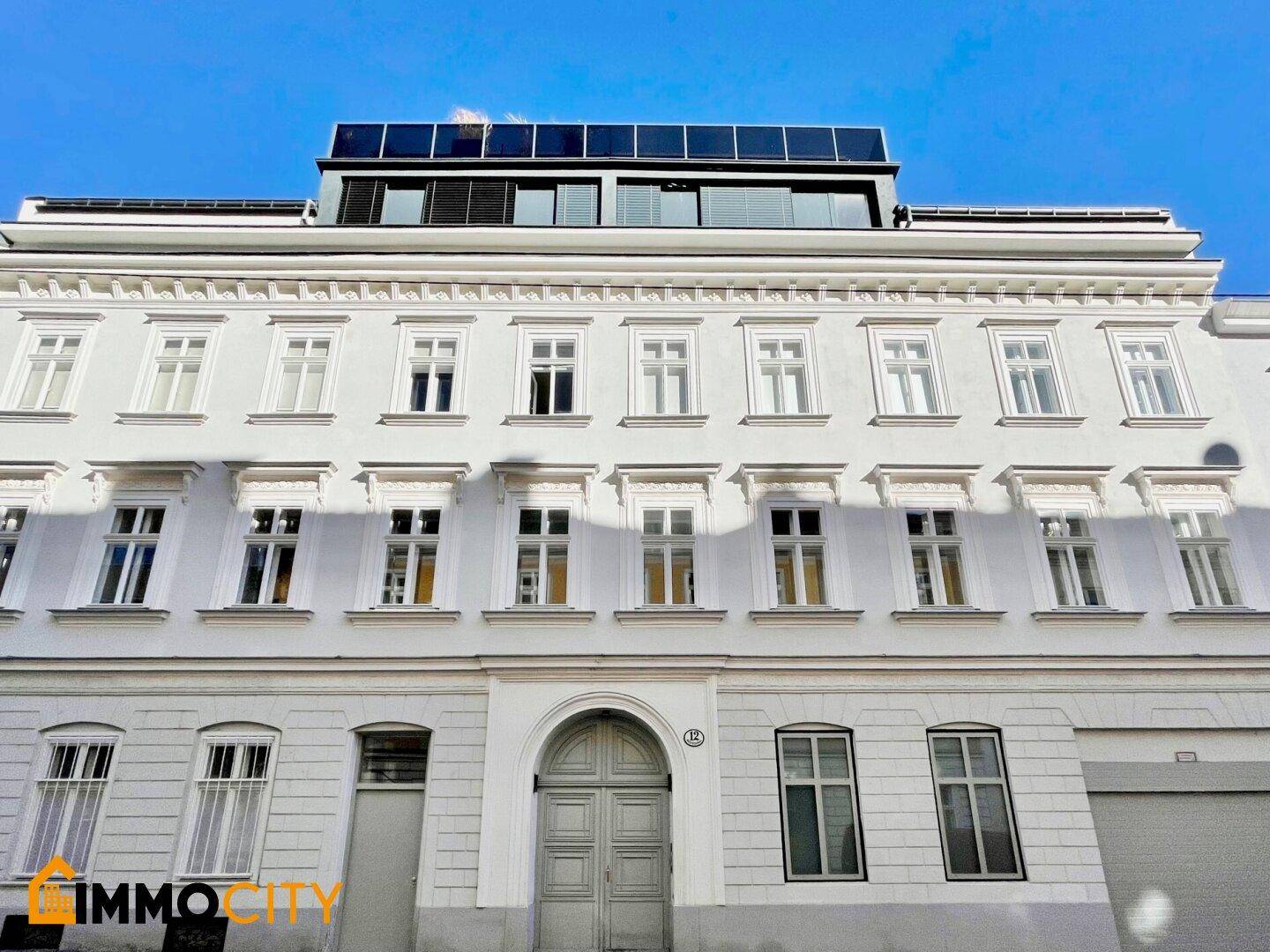 Gesamtansicht eines historischen Gebäudes mit mehreren Etagen und einer modernen Dachaufstockung mit Photovoltaikanlage.