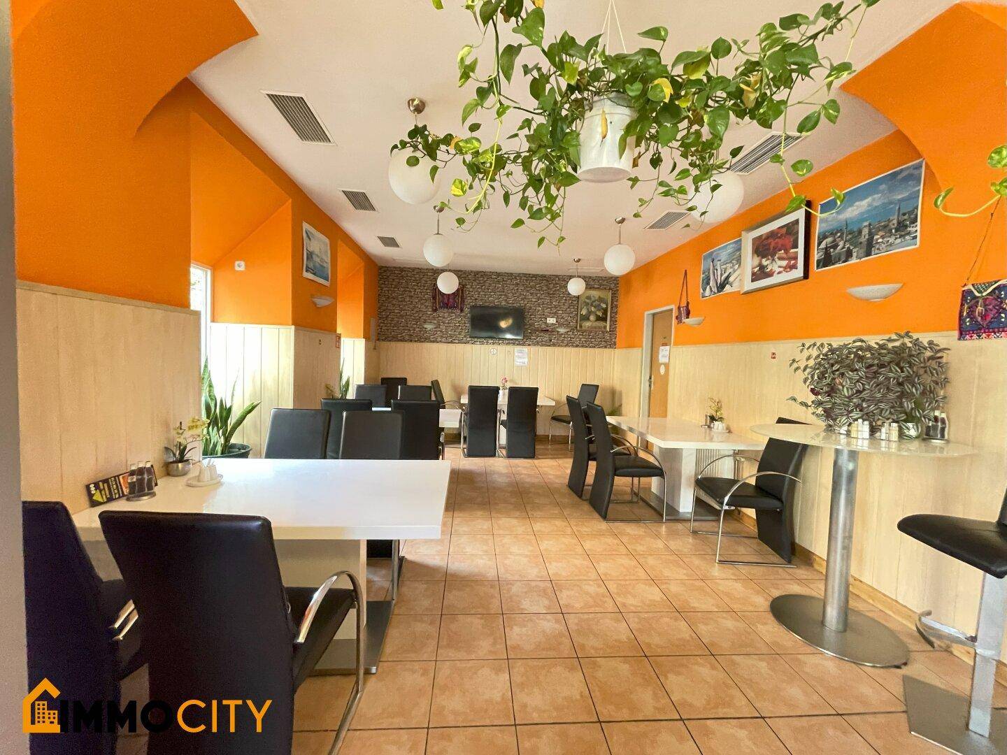 Gemütlicher Restaurantraum mit orangefarbenen Akzentwänden, hellen Tischen und dunklen Stühlen, dekoriert mit Pflanzen.