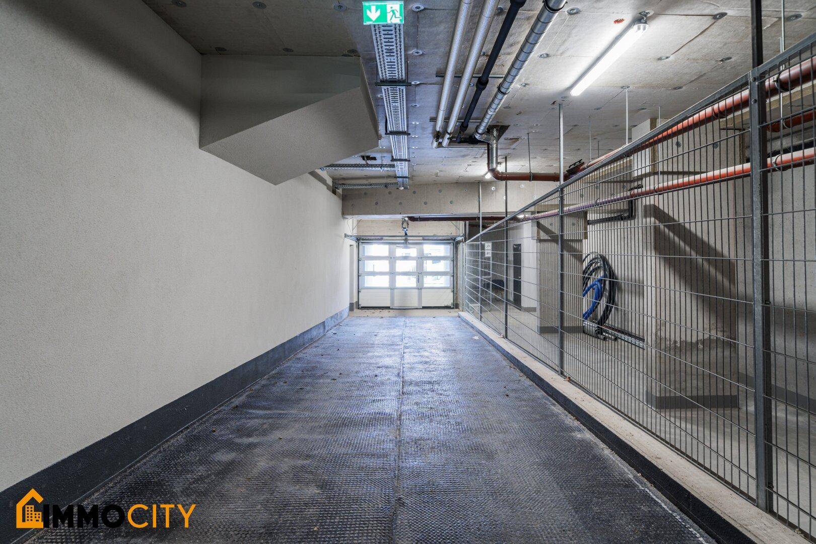Geräumige Tiefgarage mit Betonboden, Gitterabtrennungen und sichtbaren Installationen an der Decke.