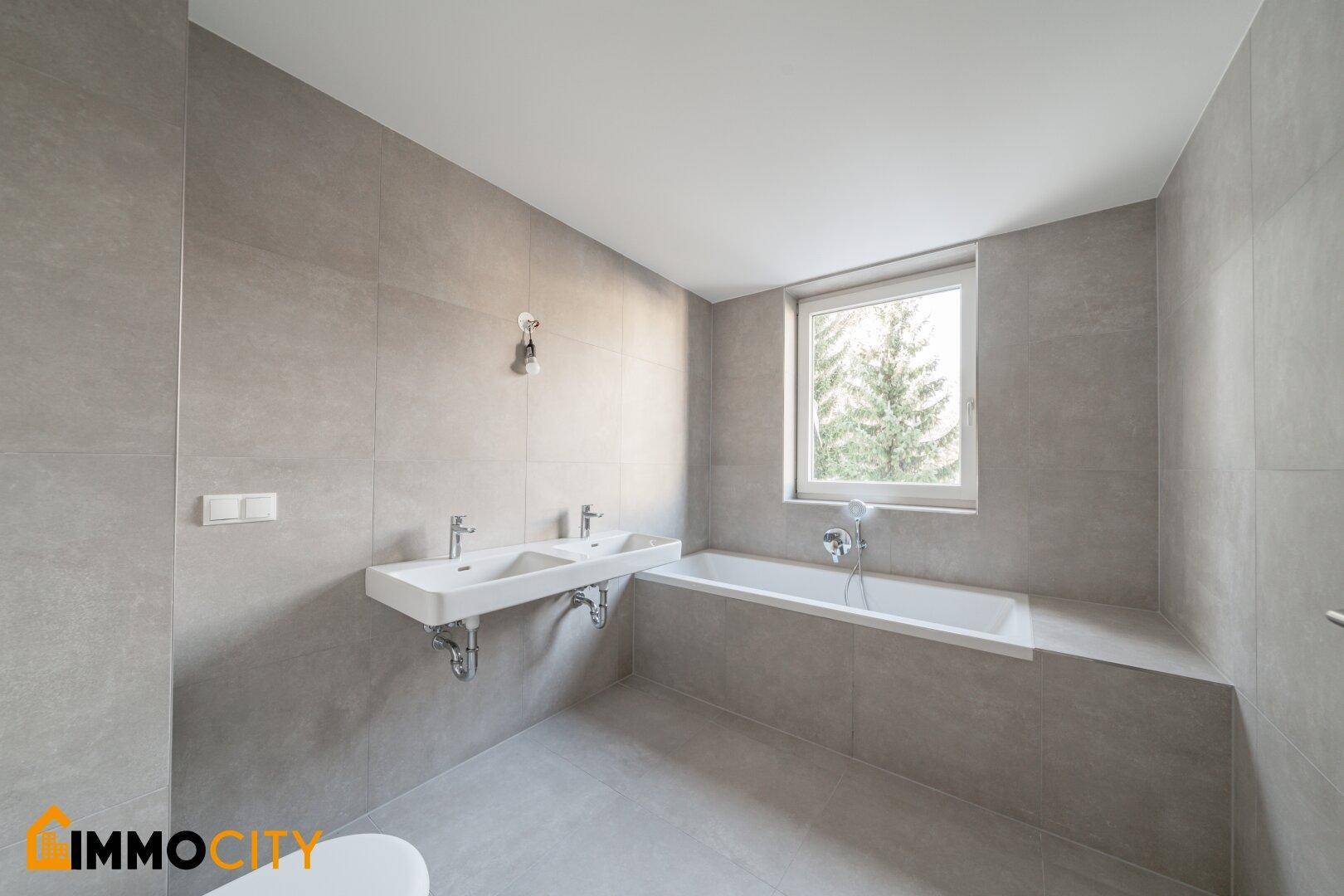 Modernes Badezimmer mit grauen Fliesen, Doppelwaschbecken und einer Badewanne unter dem Fenster.