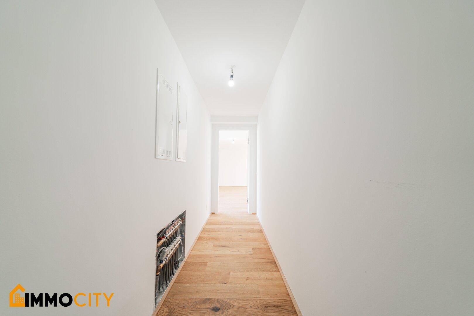 Langer, heller Flur mit Holzboden und sichtbaren Installationen an der Wand.