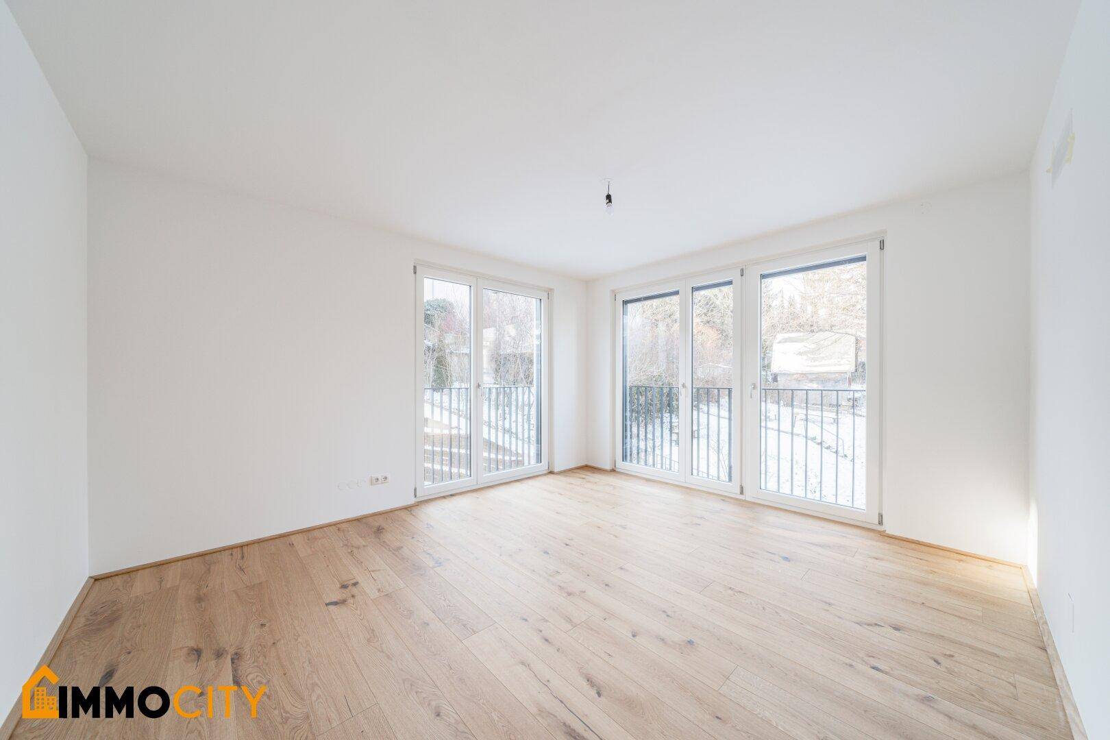 Leerer, heller Raum mit Holzboden und mehreren Fenstern, die den Blick nach draußen freigeben.