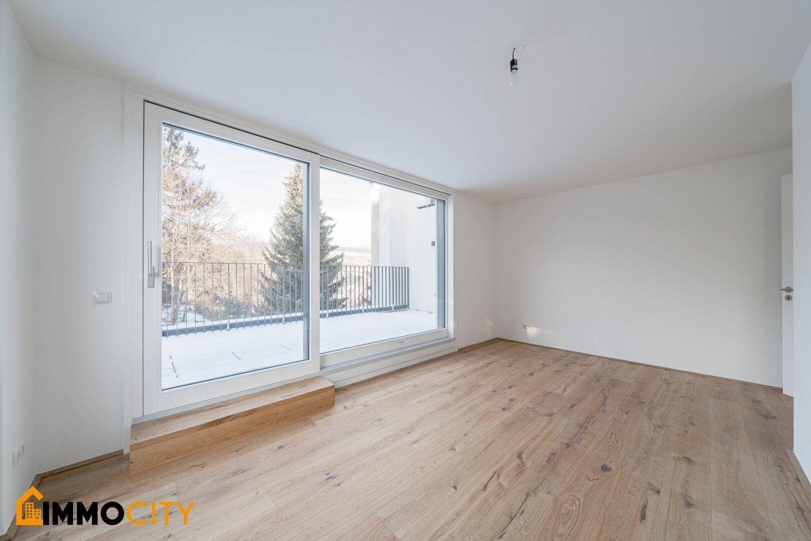 Geräumiger Raum mit Holzboden und großen Fenstern, die Zugang zu einem Balkon bieten.