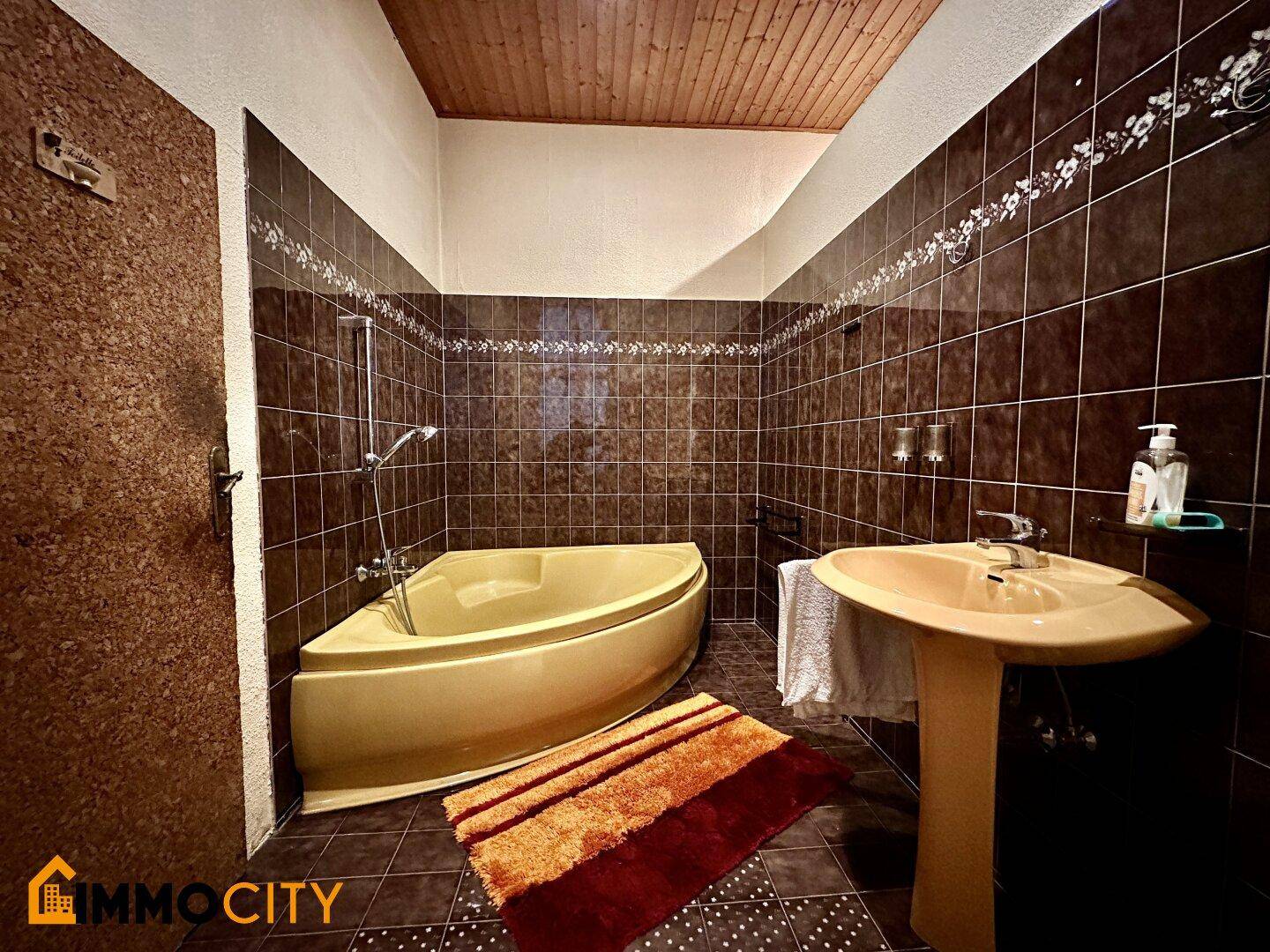 Badezimmer mit braunen Fliesen, Eckbadewanne und passendem Waschbecken, mit Holzdecke.
