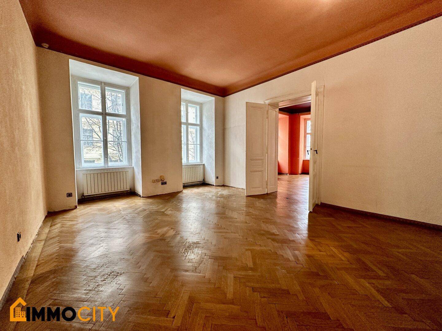 Geräumiges Zimmer mit Fischgrätparkett, hohen Decken und Blick in angrenzende Räume.