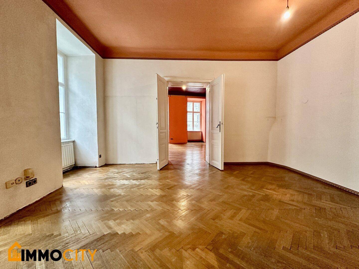 Geräumiges Zimmer mit Fischgrätparkett, hohen Decken und Blick in angrenzende Räume.