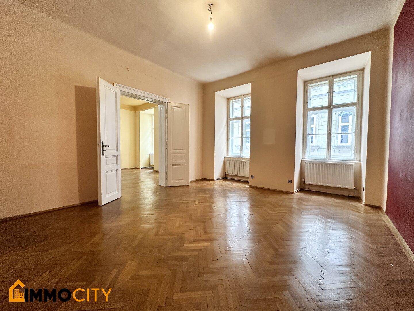 Geräumiges Zimmer mit Fischgrätparkett und hohen Decken, mit Blick in angrenzende Räume.