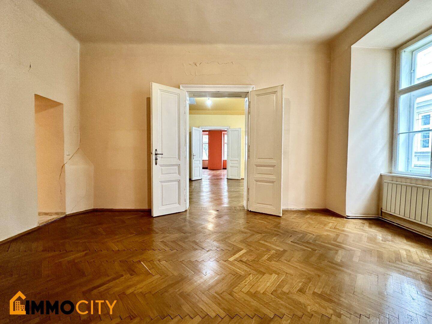 Helles Zimmer mit Fischgrätparkett und hohen Decken, mit Blick durch offene Türen in weitere Räume.