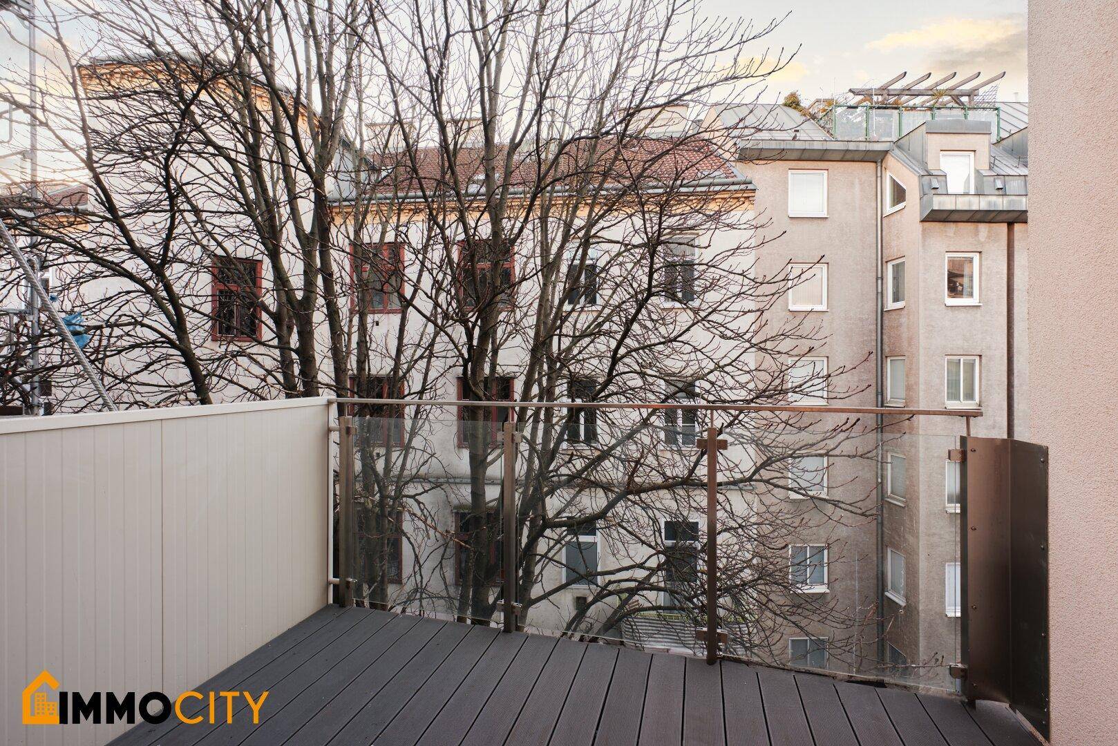 Moderner Balkon mit Glasgeländer und Blick auf die umliegenden Gebäude und Bäume.